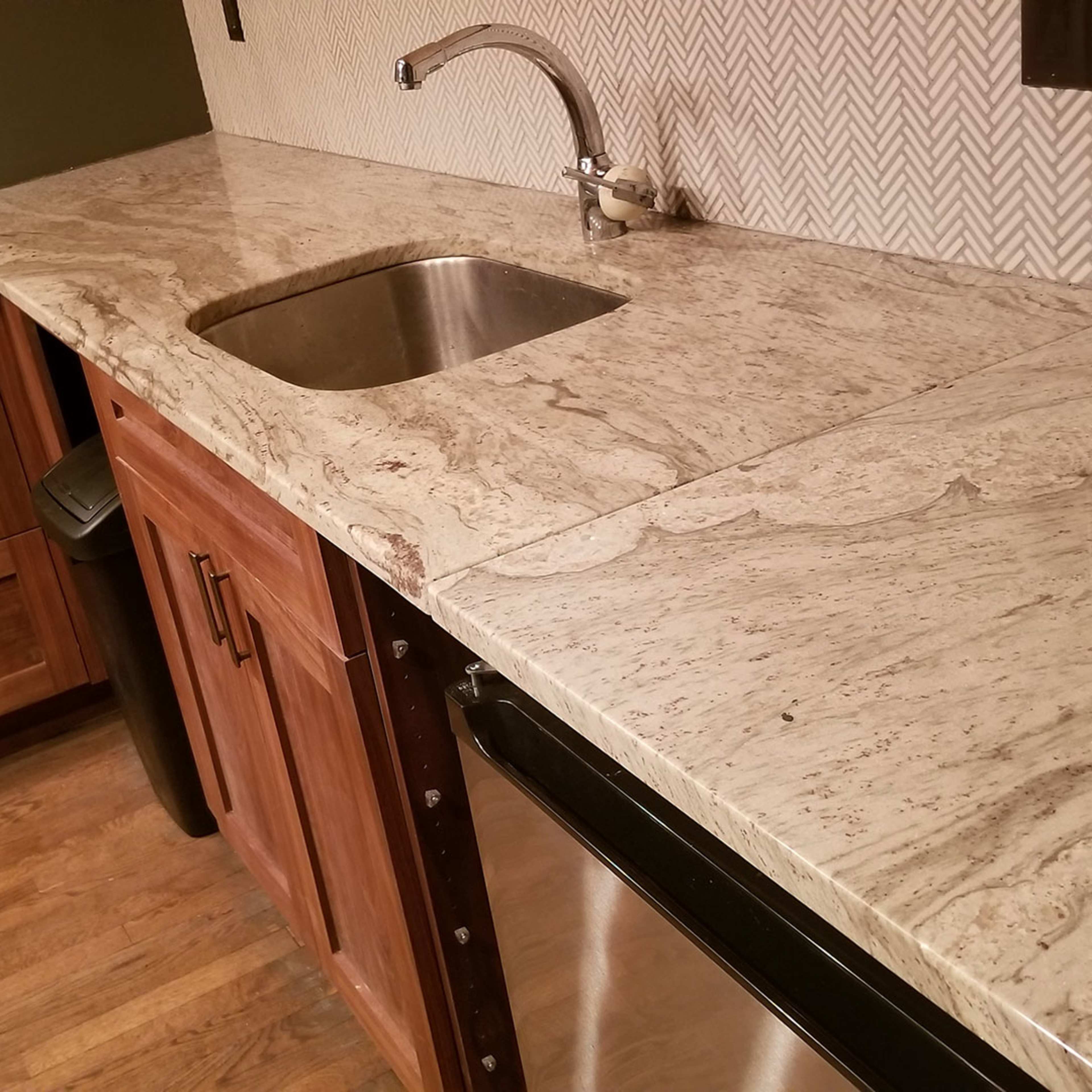 A kitchen corner featuring wooden cabinetry, a stainless steel sink, and a granite countertop with a herringbone-patterned backsplash.
