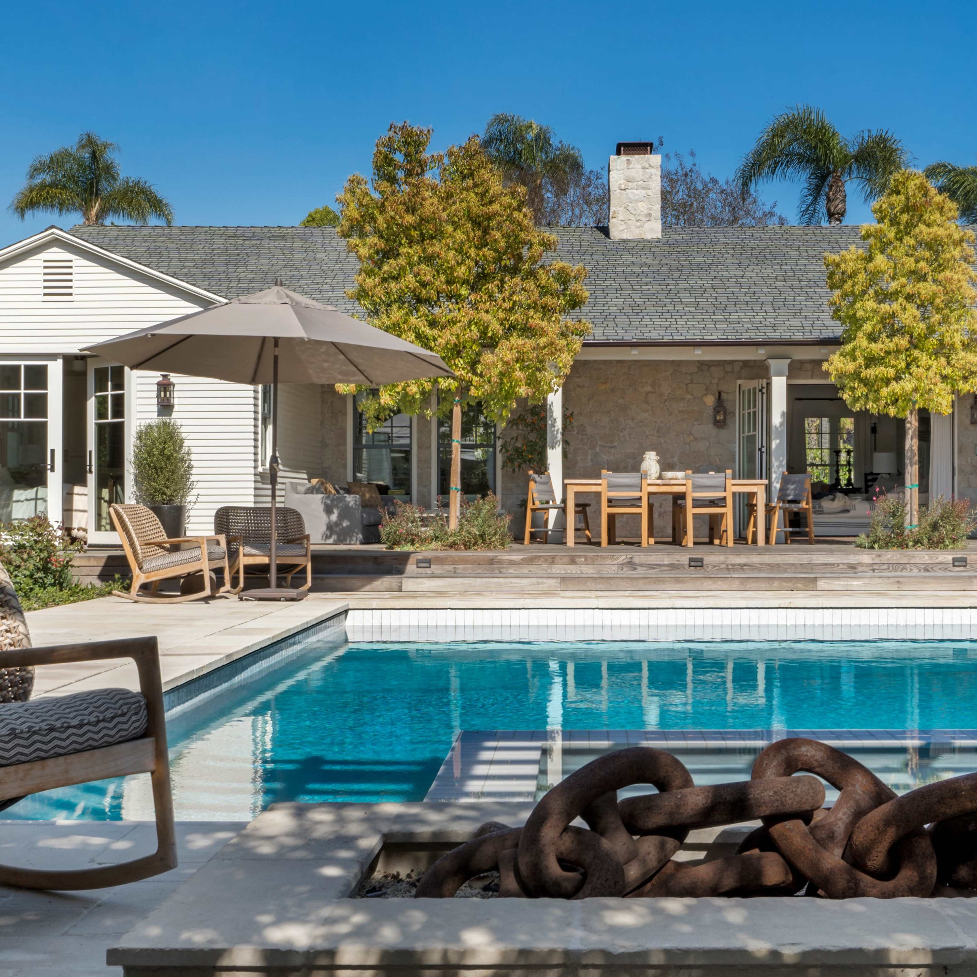 A modern backyard features a swimming pool with lounge chairs, an umbrella, and a dining area surrounded by greenery and stone architecture.