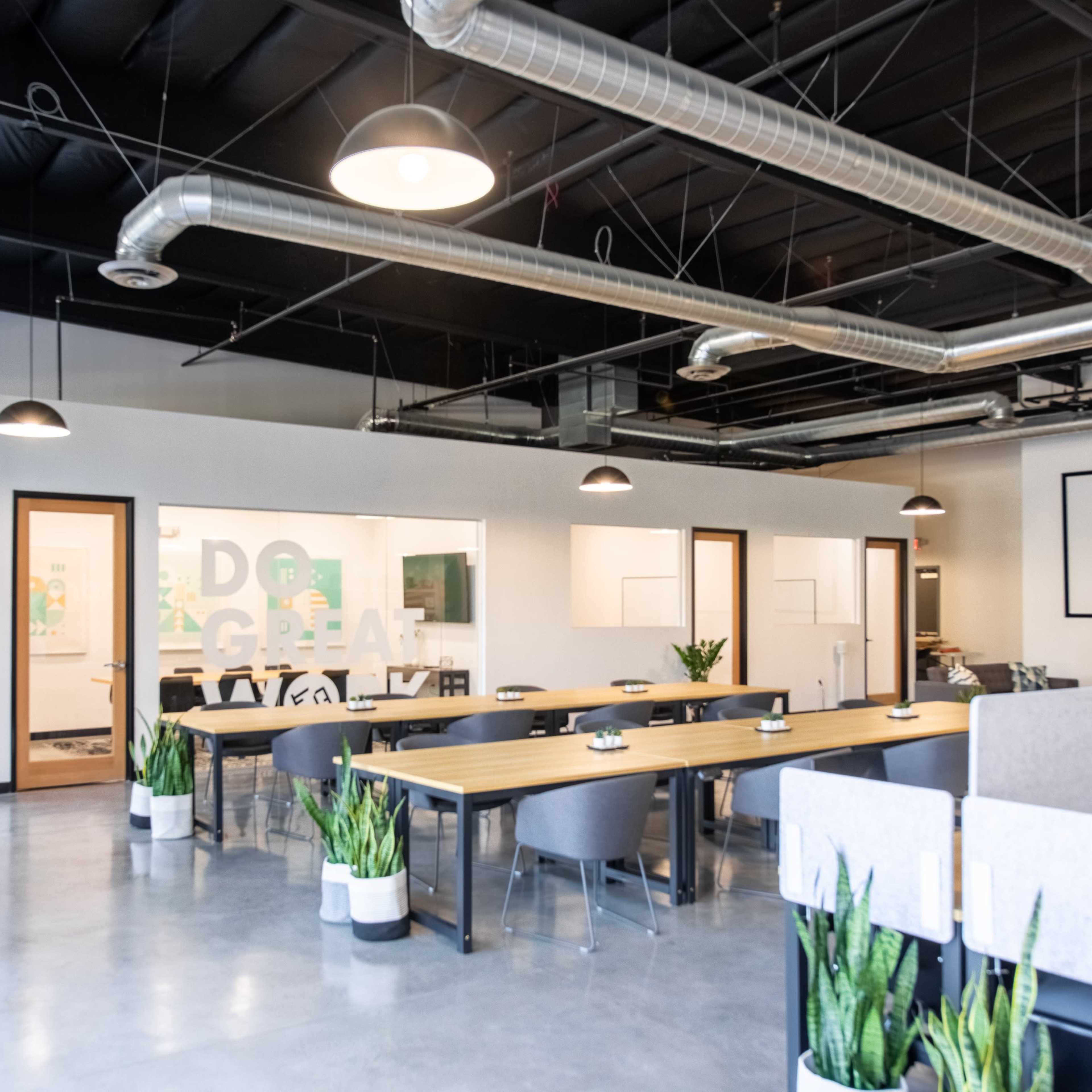 A modern, open office space featuring desks arranged in a communal area, large windows, and plants in decorative pots.