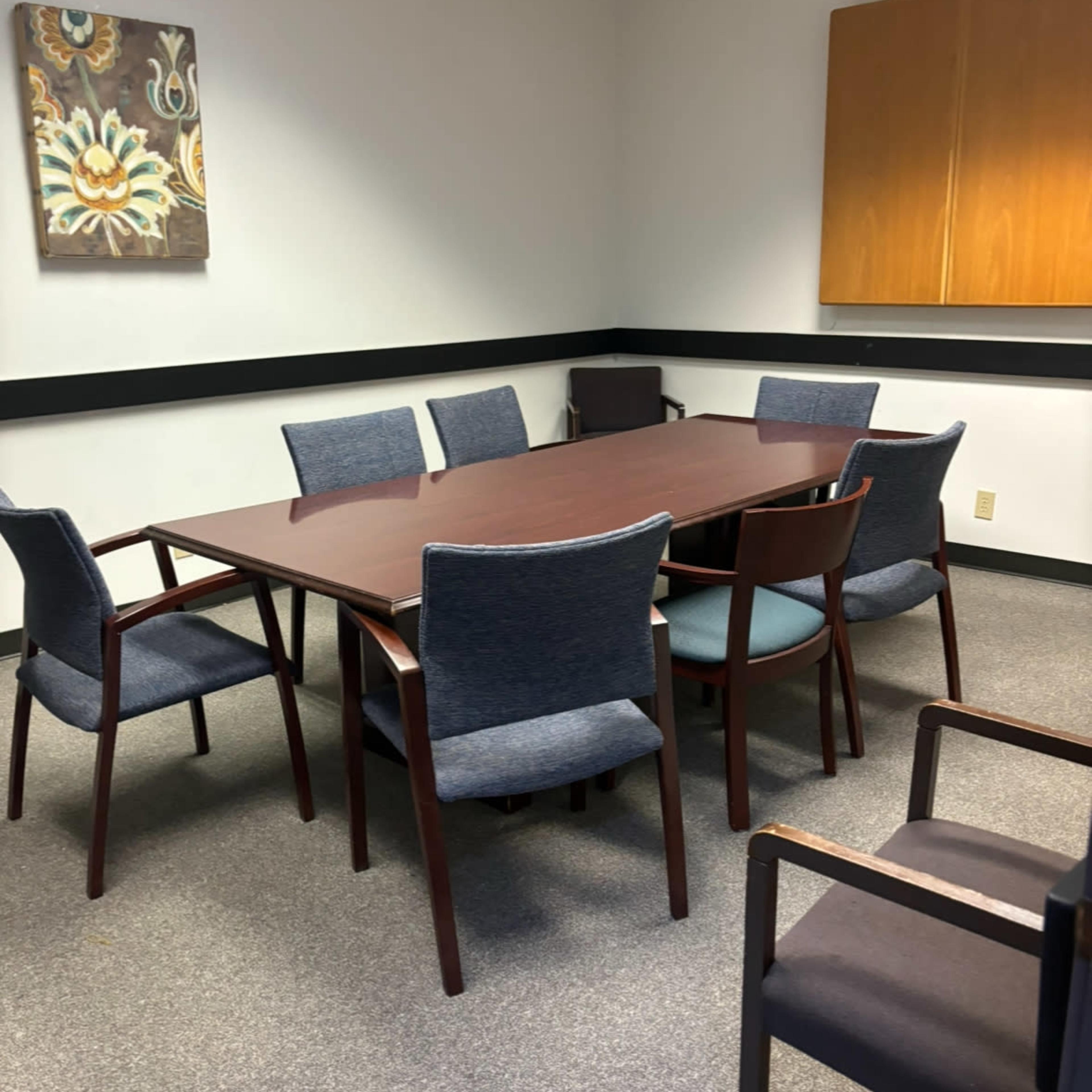 A conference room features a large wooden table surrounded by several chairs and a wall-mounted cabinet.
