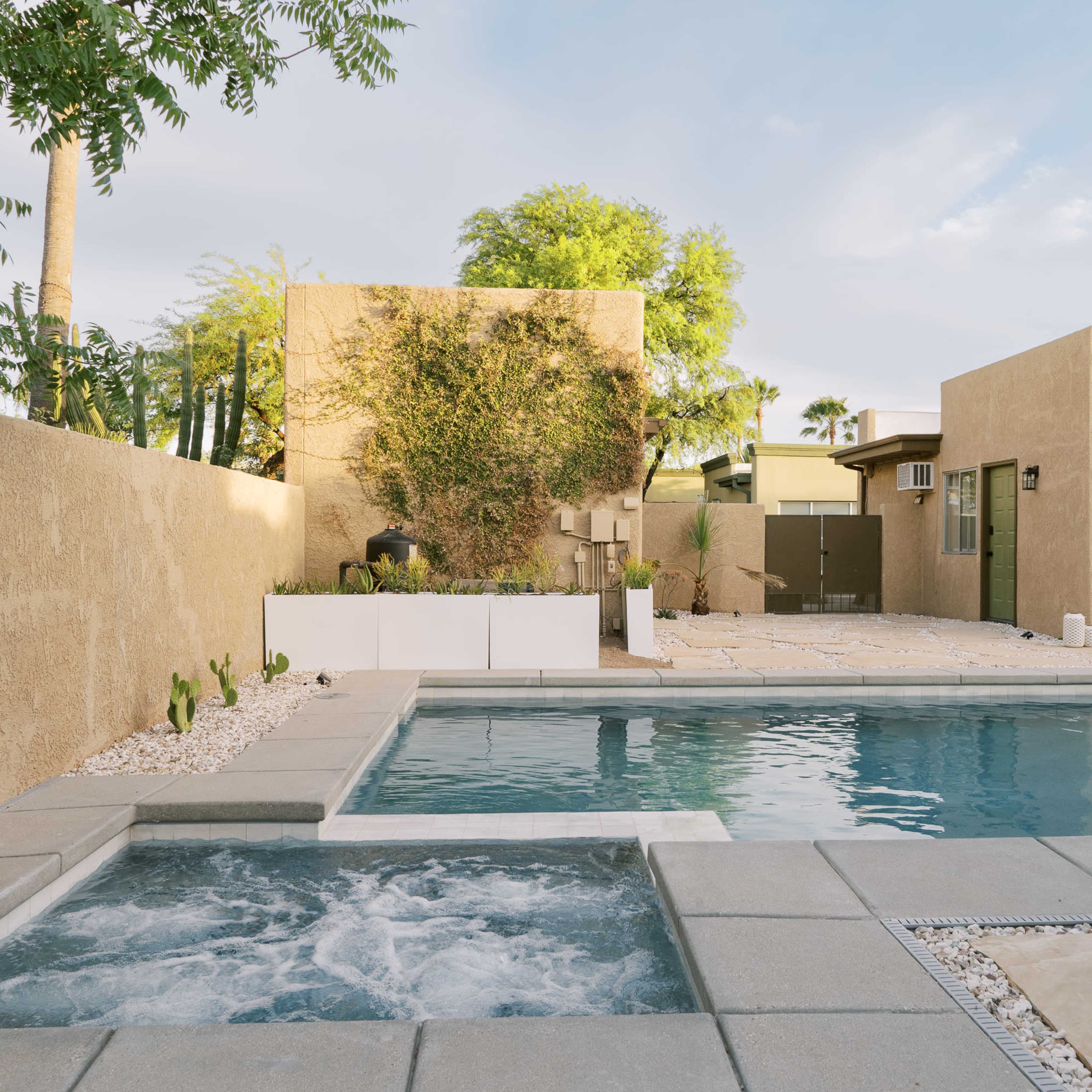 A modern backyard with a swimming pool and spa, surrounded by beige stucco walls and landscaped areas.