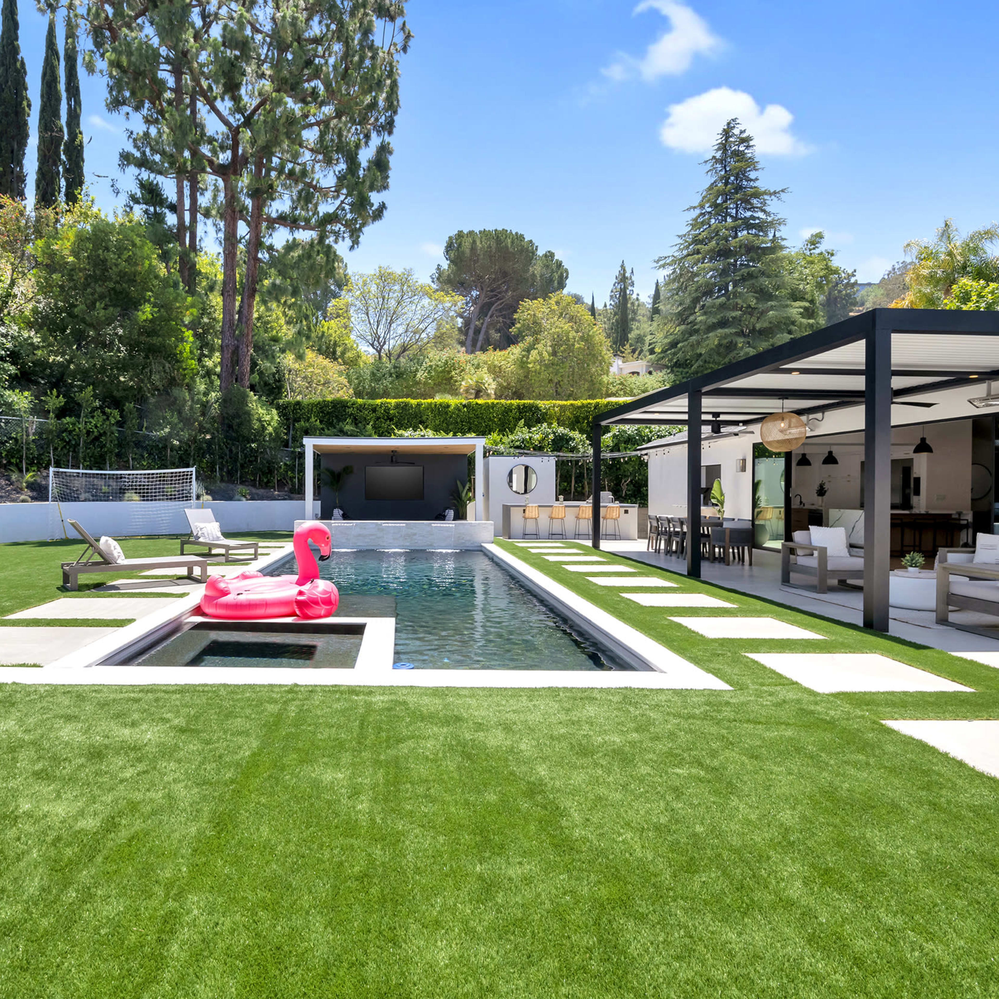 A landscaped backyard featuring a swimming pool with a pink flamingo float, surrounded by manicured grass and modern patio furniture.