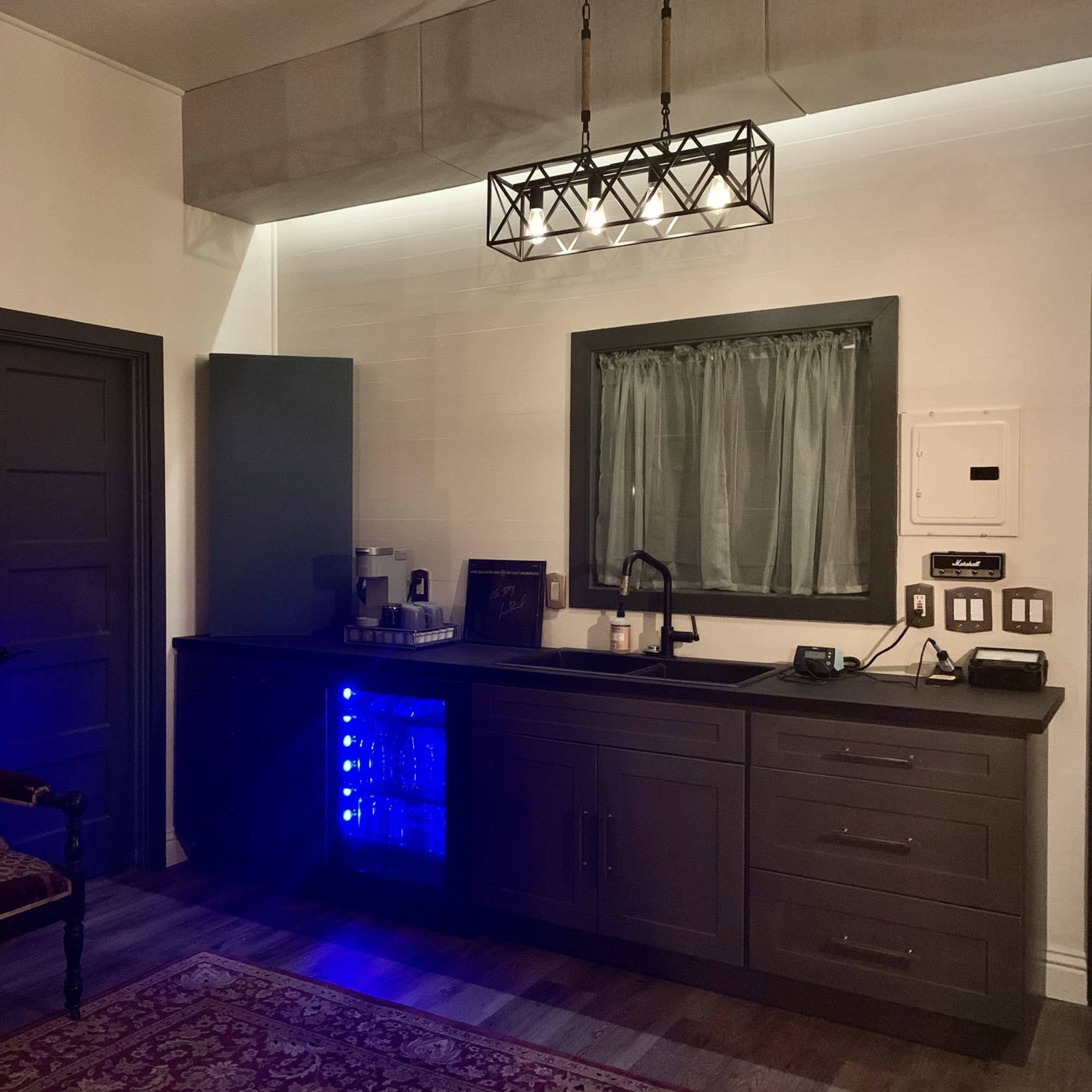 A kitchen area featuring a dark cabinet with a blue-lit mini refrigerator, a sink, and a vintage-style chair beside it.