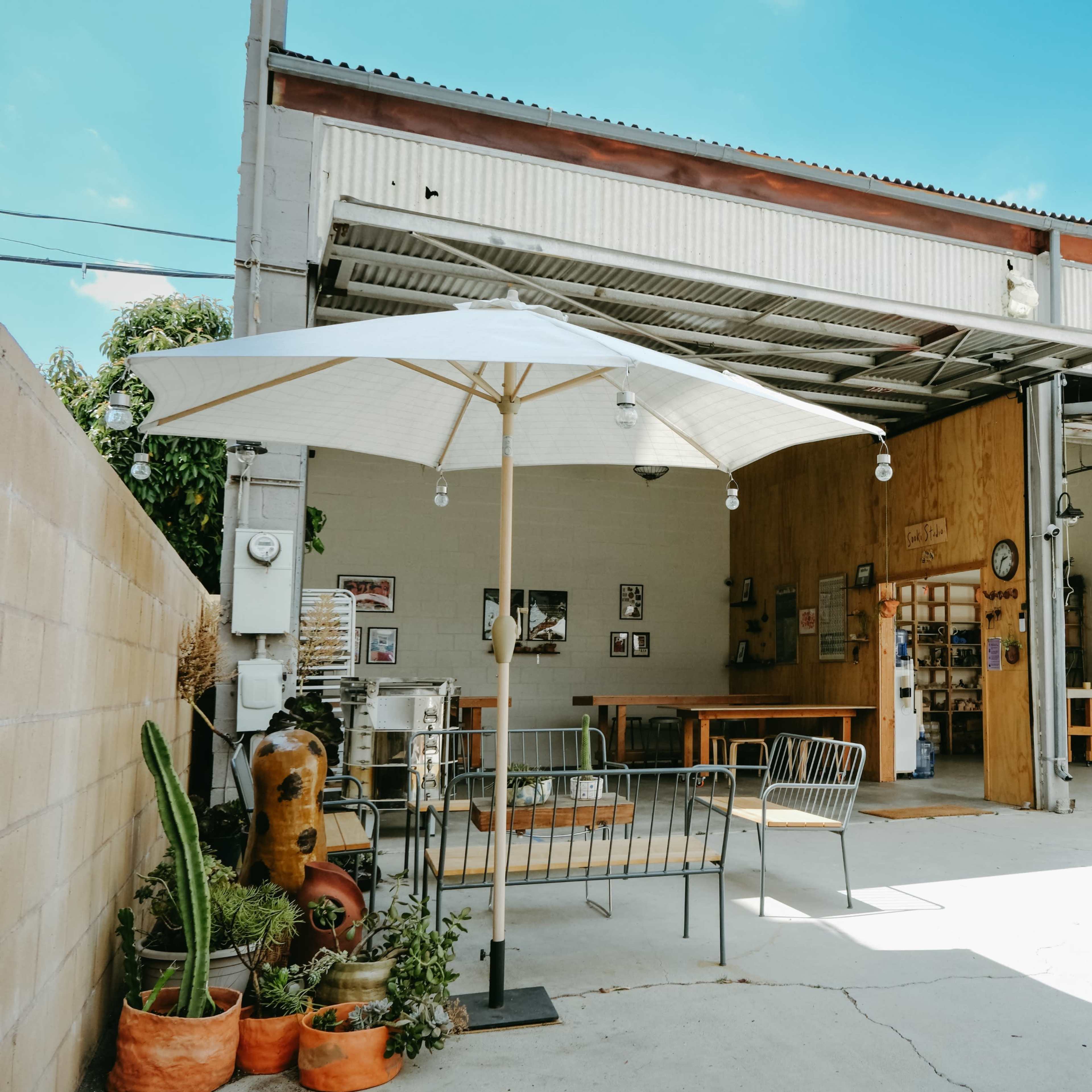 A spacious outdoor area of a café or workshop with a large umbrella, seating, and a few tables under a well-lit, open garage-style structure.