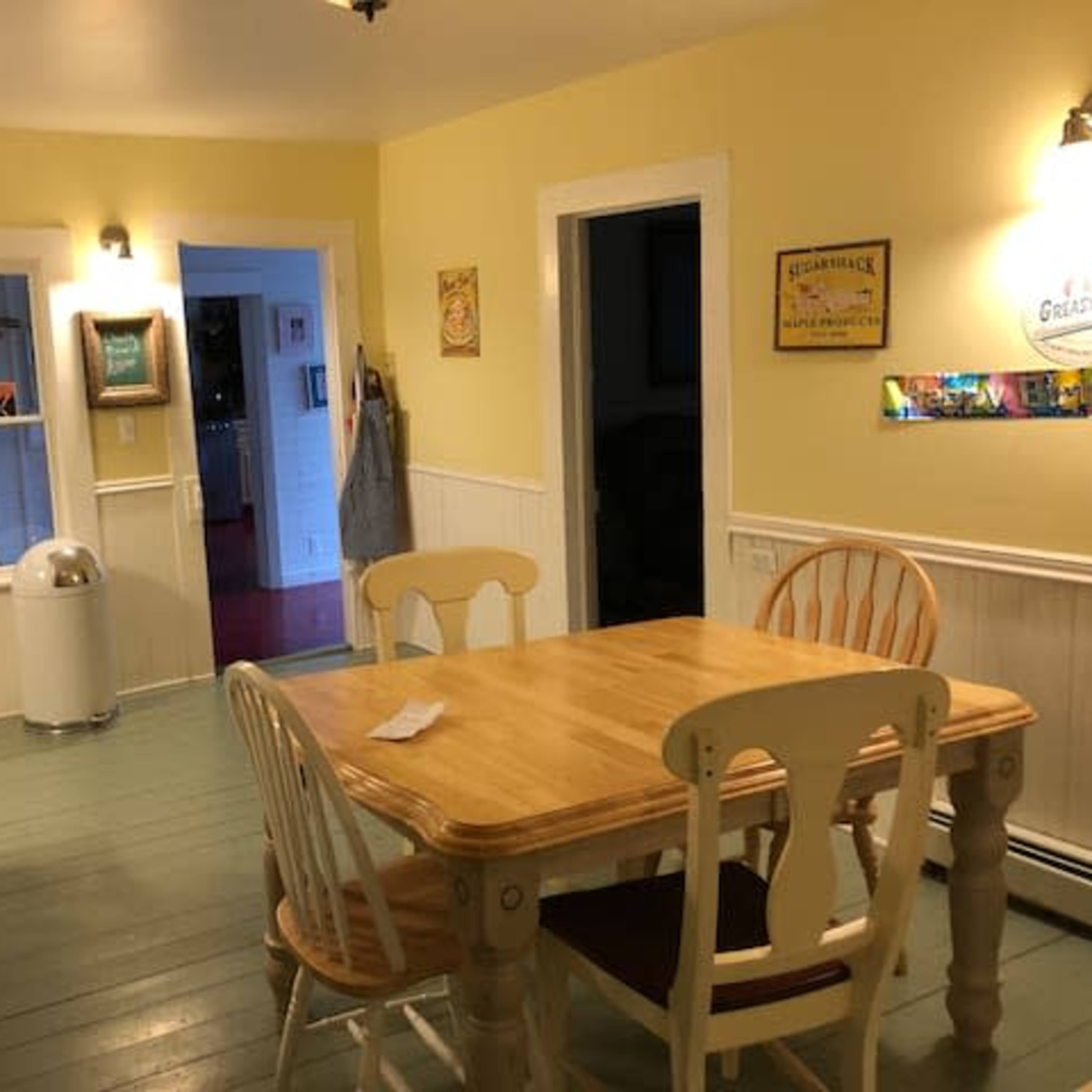 A bright yellow kitchen with a wooden table and chairs, a doorway leading to another room, and various decorations on the walls.