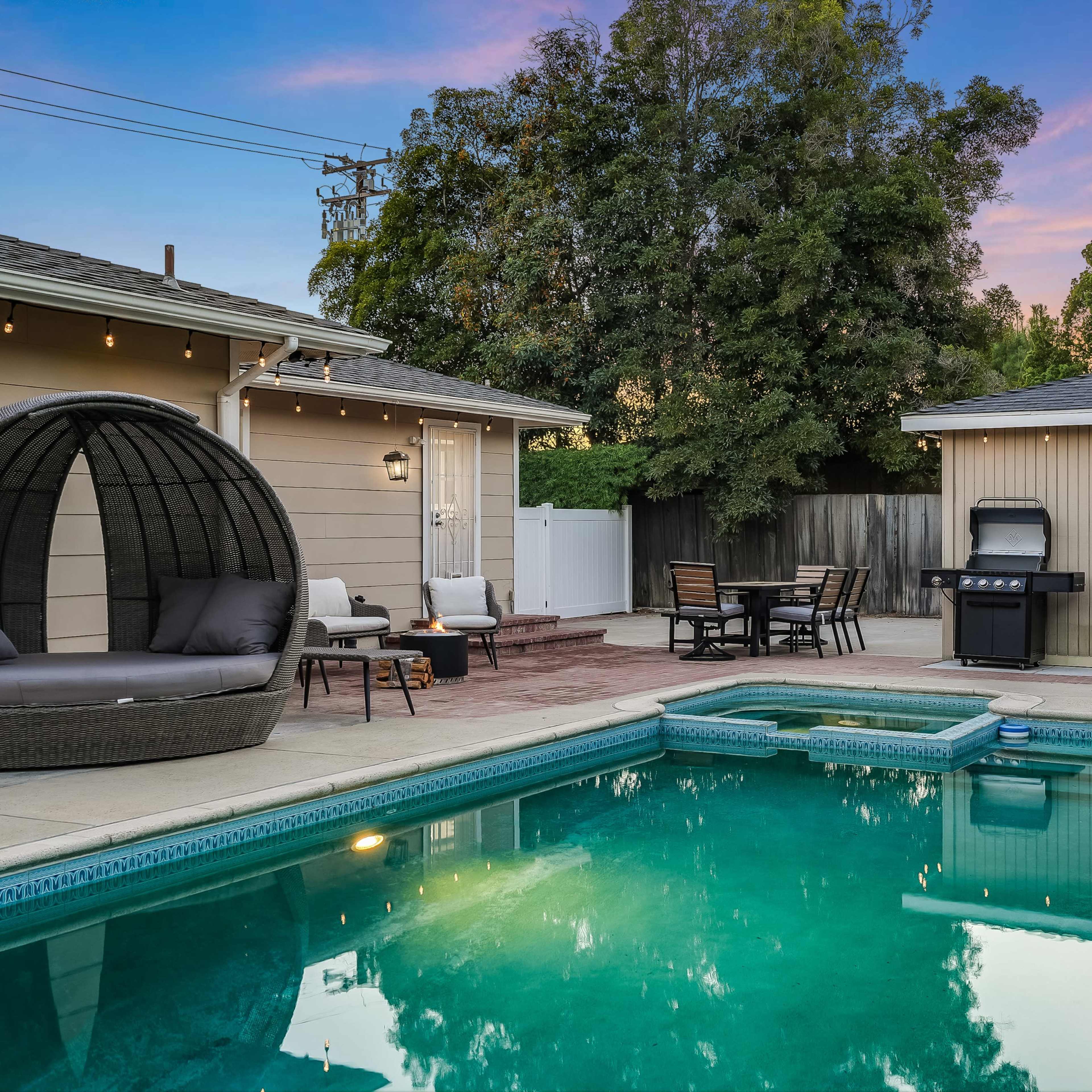 A backyard with a swimming pool, outdoor furniture, and a barbecue grill surrounded by trees and a house.