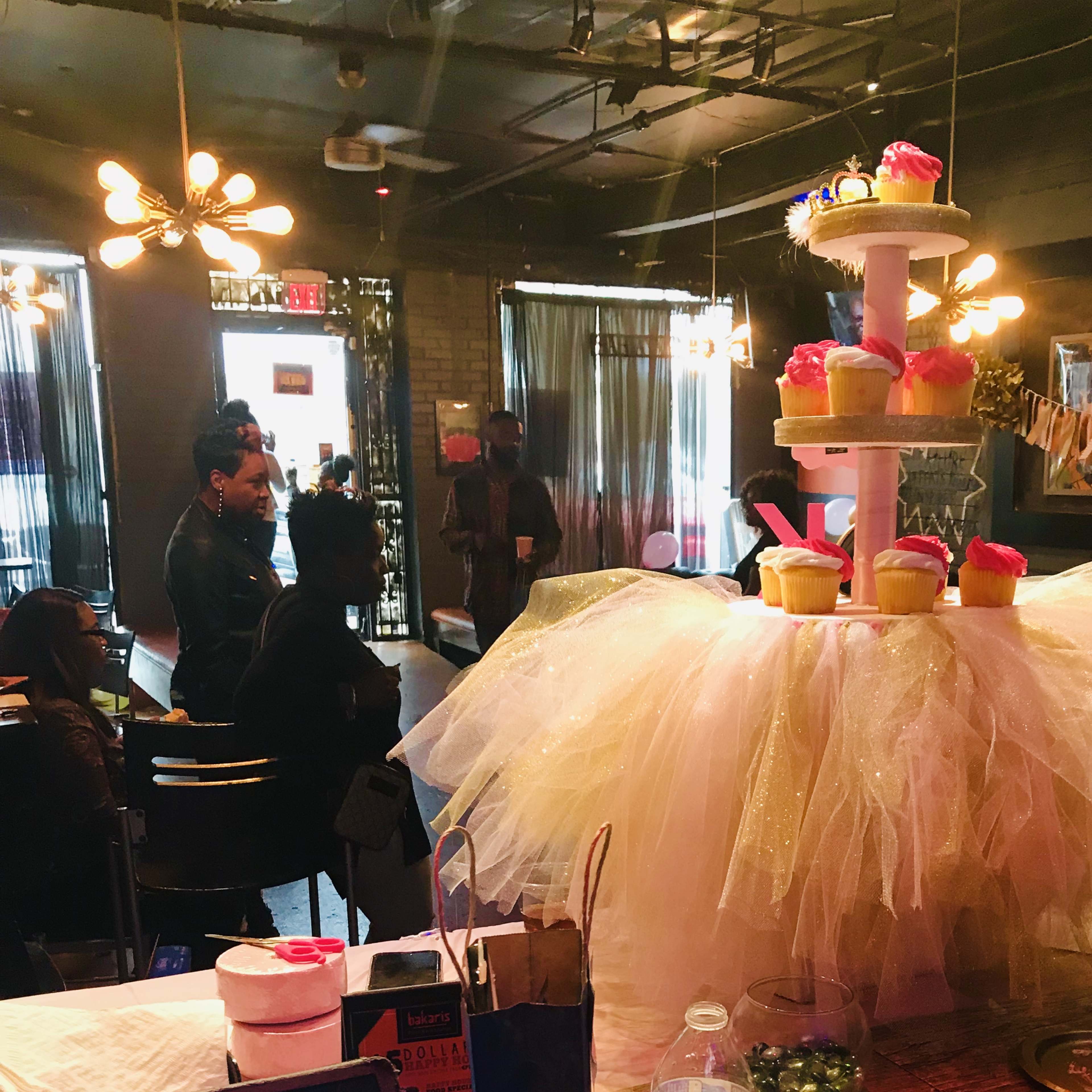 A large, tiered cupcake display with pink frosting and a tulle skirt is the centerpiece in a decorated café where people are mingling.