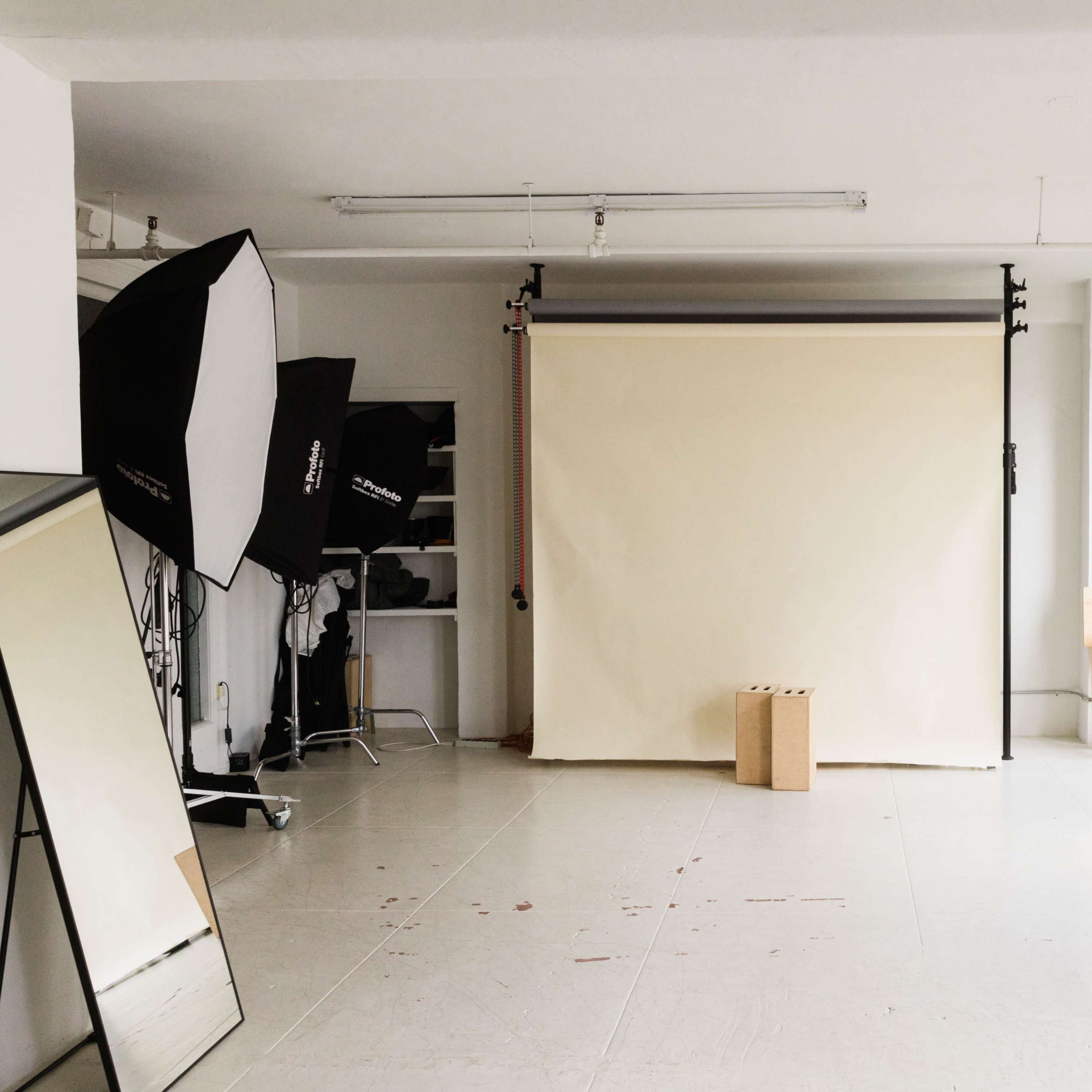 A photography studio features a beige backdrop, two large softbox lights, a mirror, and a wooden stool situated in a well-lit space with large windows.