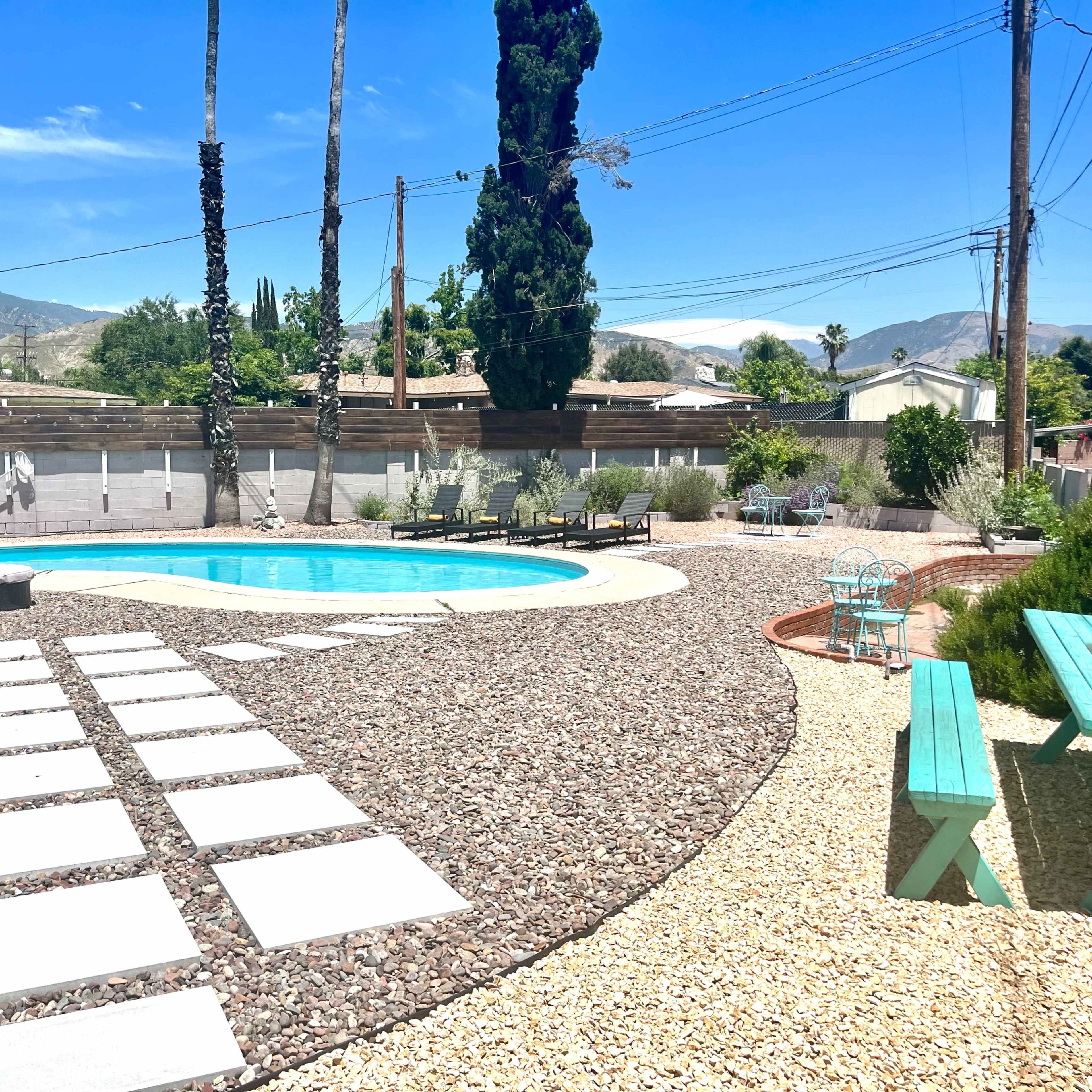 A backyard with a swimming pool surrounded by stone pathways, patio furniture, and gravel landscaping with mountains in the background.