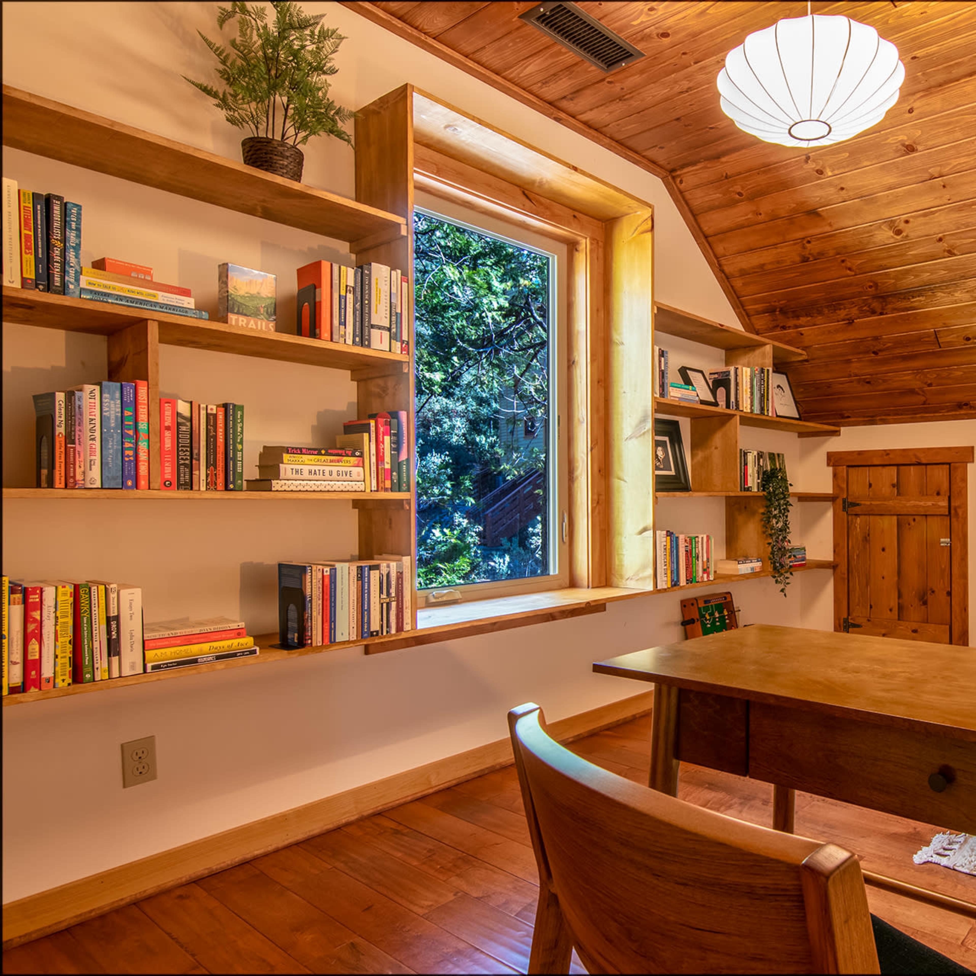 A cozy room with wooden walls and ceiling features a desk, bookshelves filled with books, and a large window overlooking greenery.