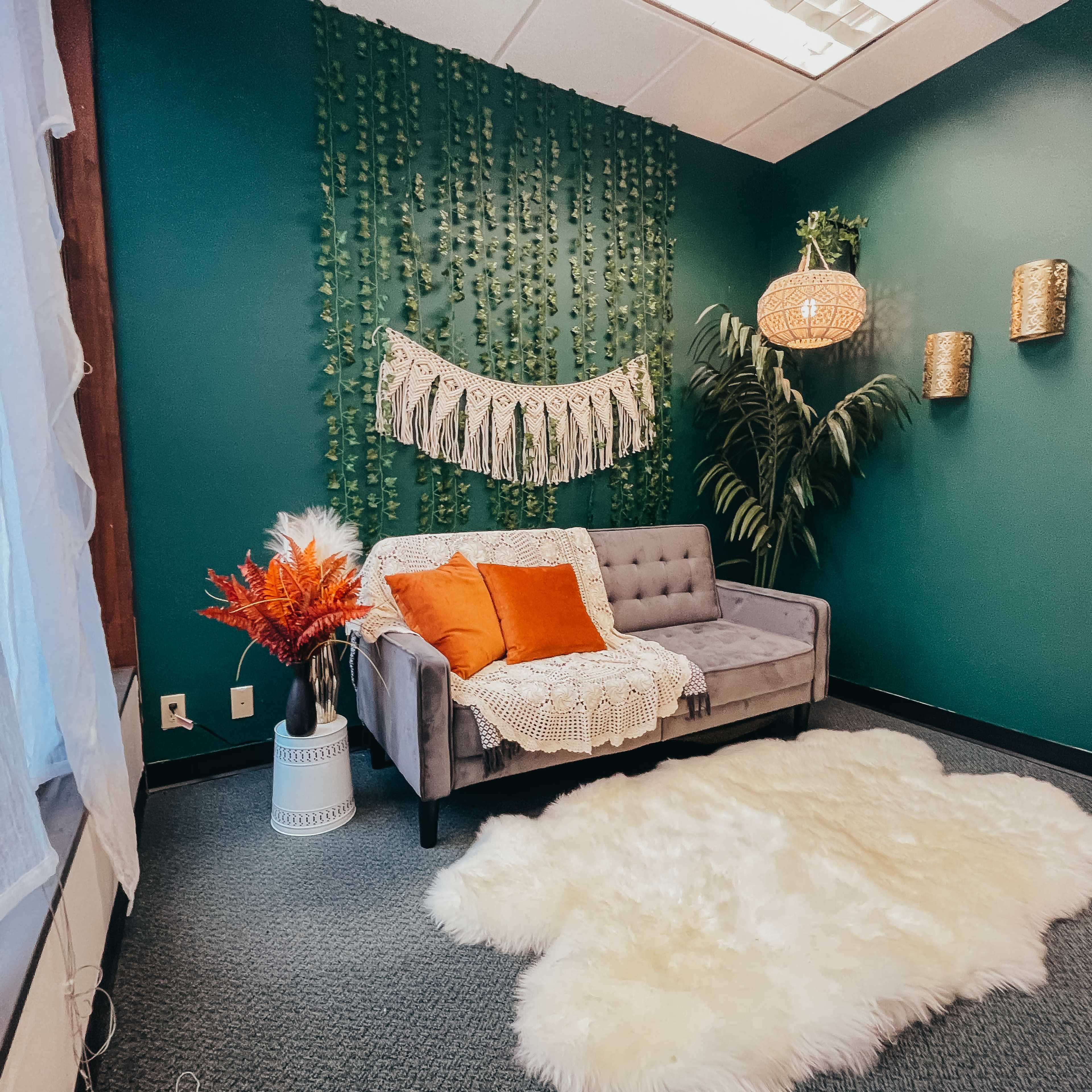 A cozy room featuring a gray couch adorned with orange pillows, a decorative wall hanging, and greenery, alongside a white faux fur rug on a carpeted floor.