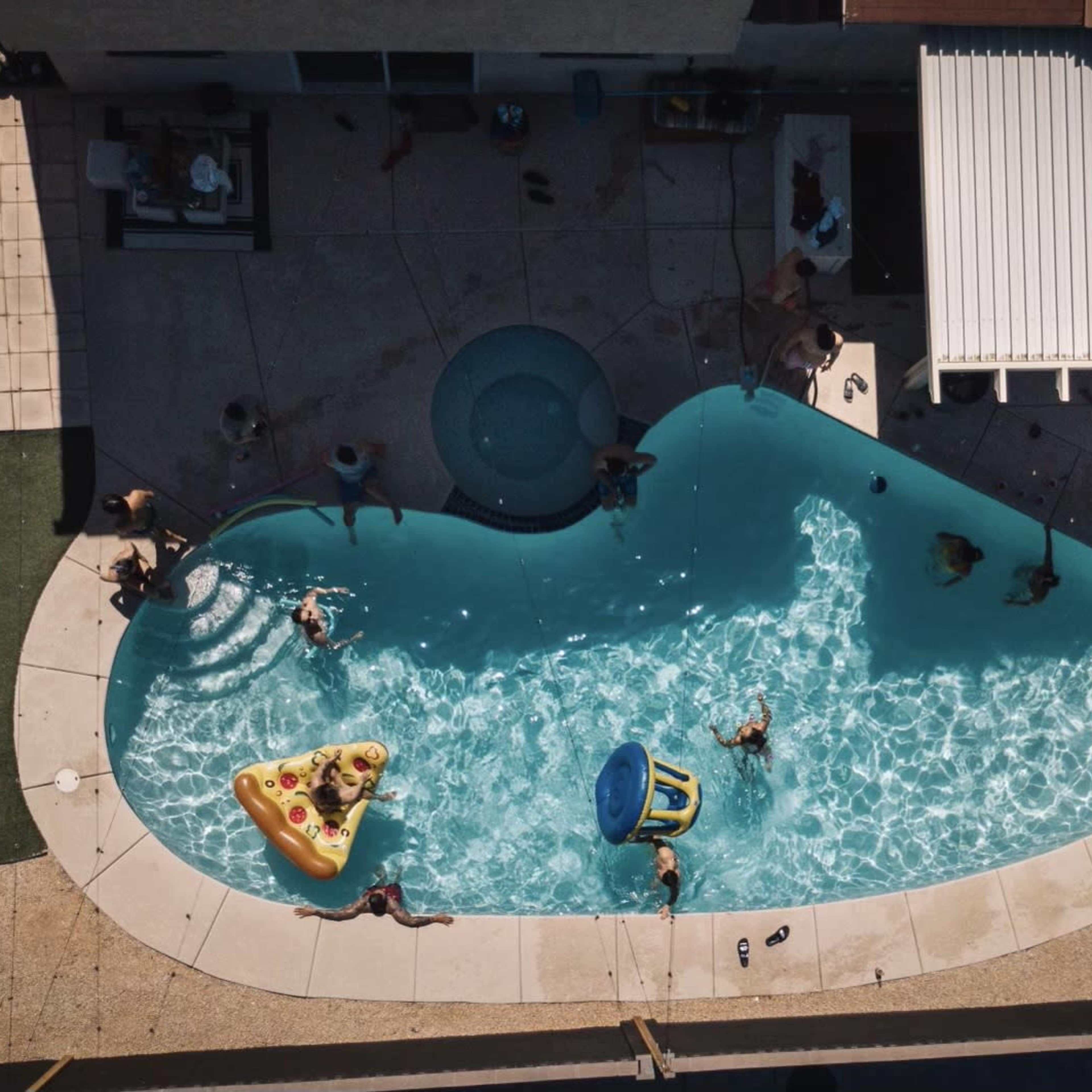 An aerial view shows a swimming pool surrounded by people enjoying various activities, with inflatable toys scattered throughout the water.