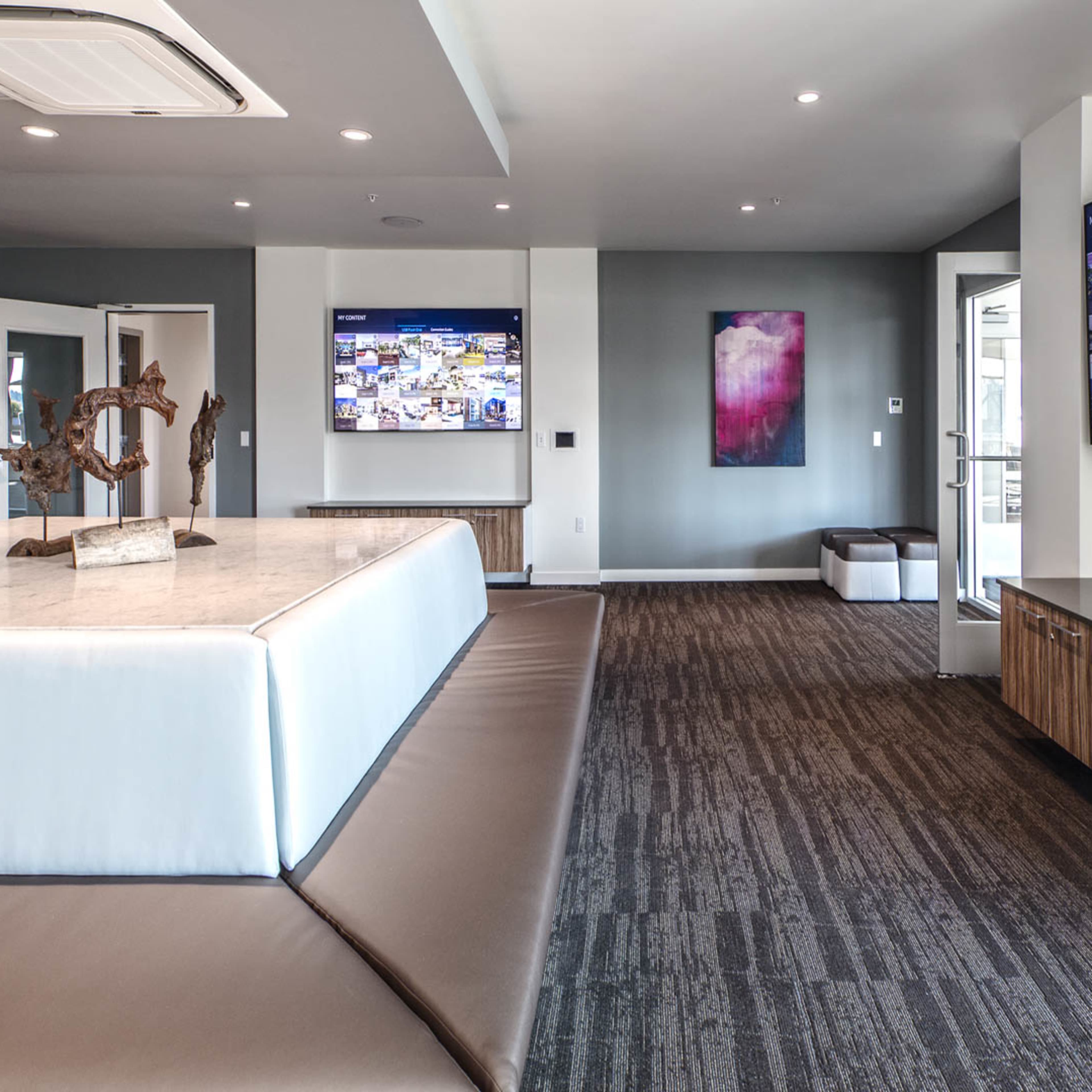 A modern interior space featuring a large central island with a marble countertop, two wall-mounted screens displaying images, and contemporary artwork on the walls.