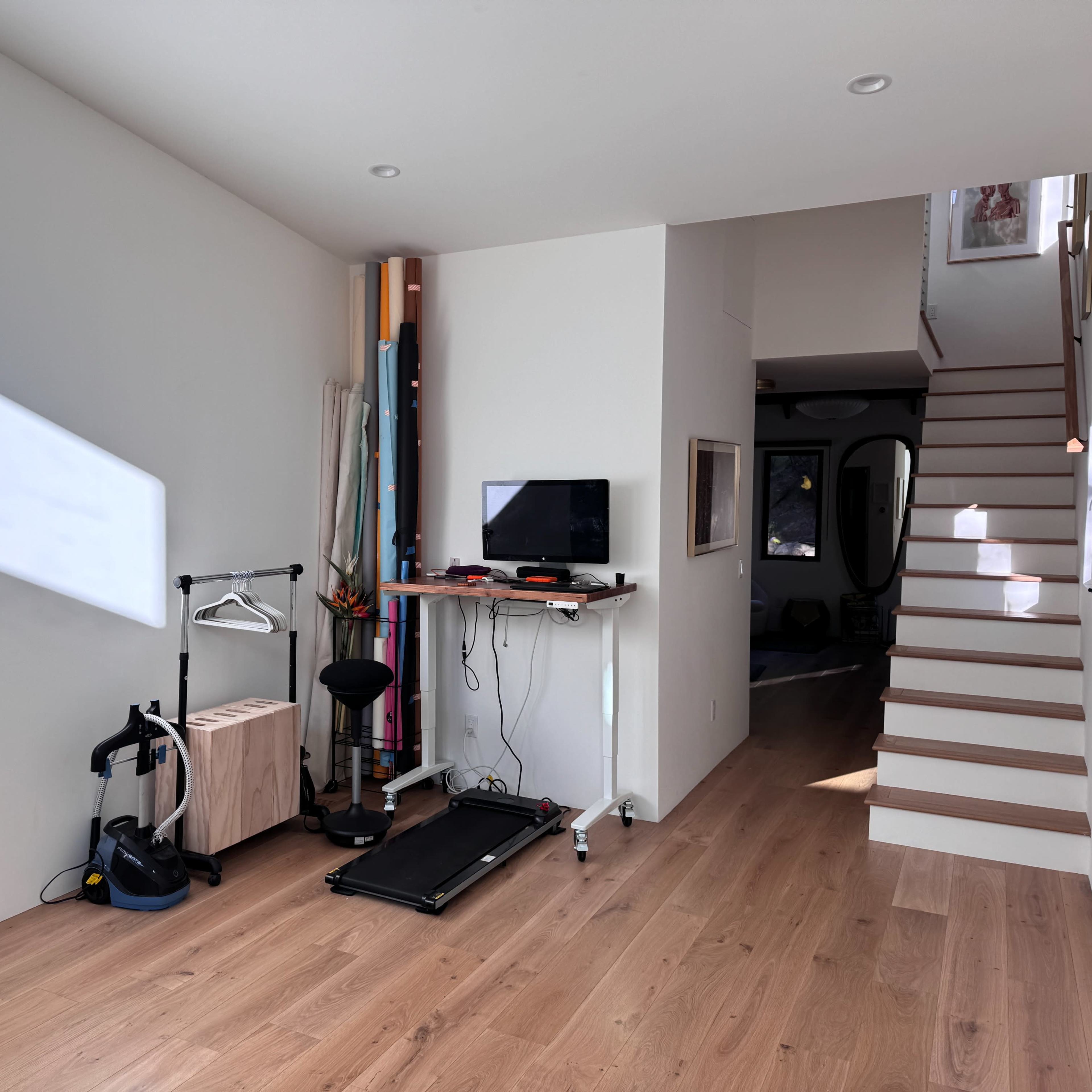 A partially furnished room features a treadmill, a desk with a monitor, and a staircase leading to another level.