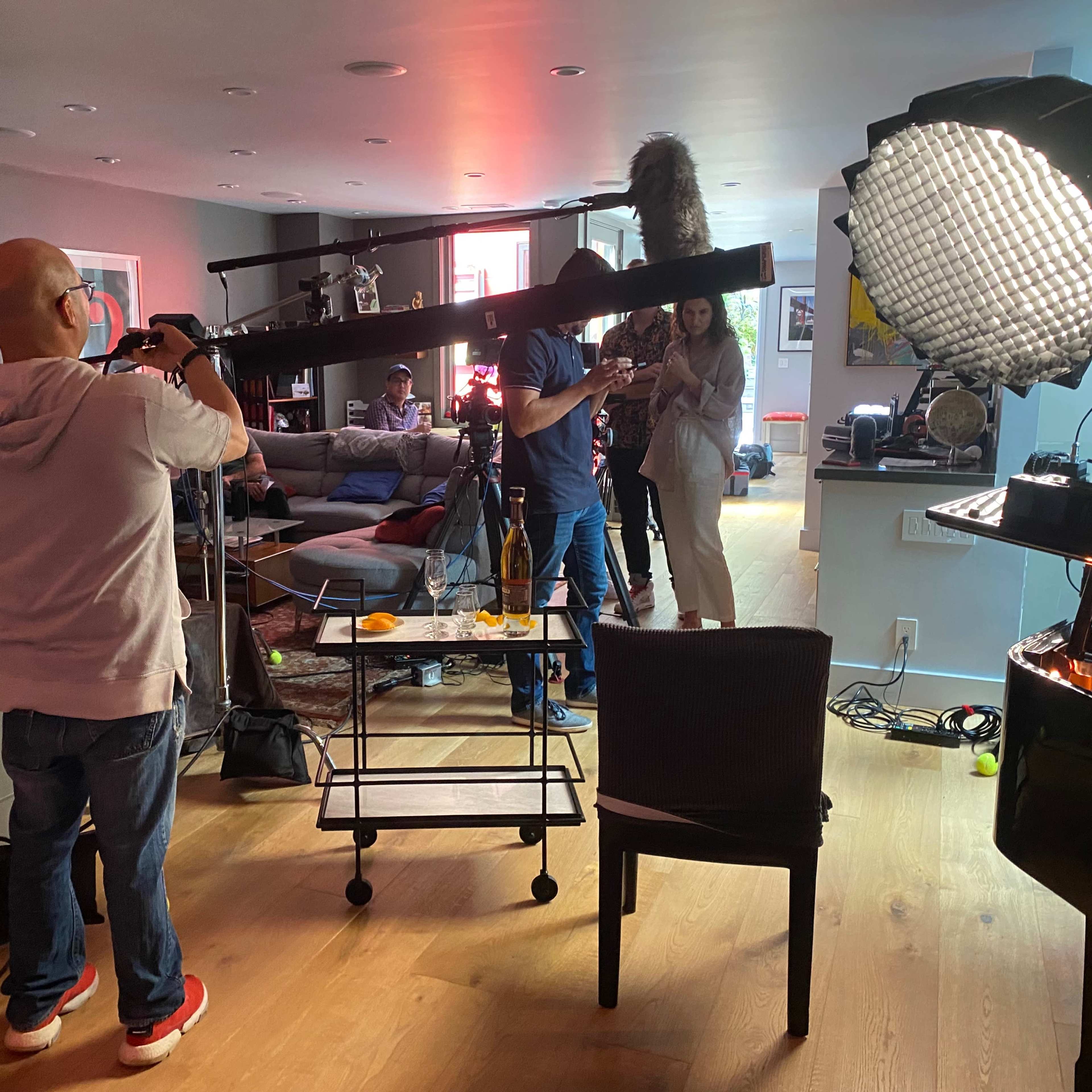 A film crew is set up in a living room, preparing equipment while one person holds a camera and another adjusts a boom microphone.