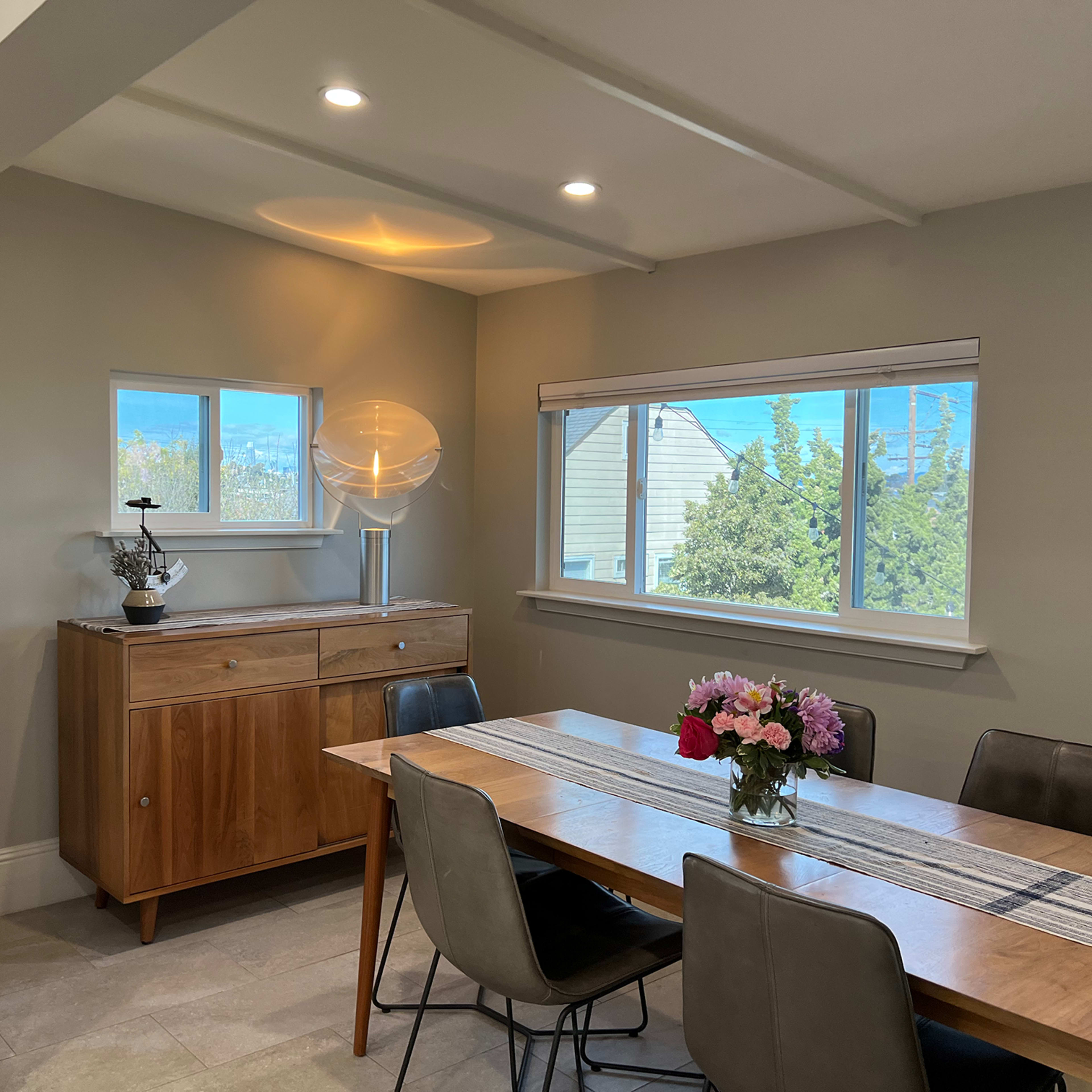 A dining area with a wooden table surrounded by chairs, a sideboard cabinet, and large windows with a view of trees outside.