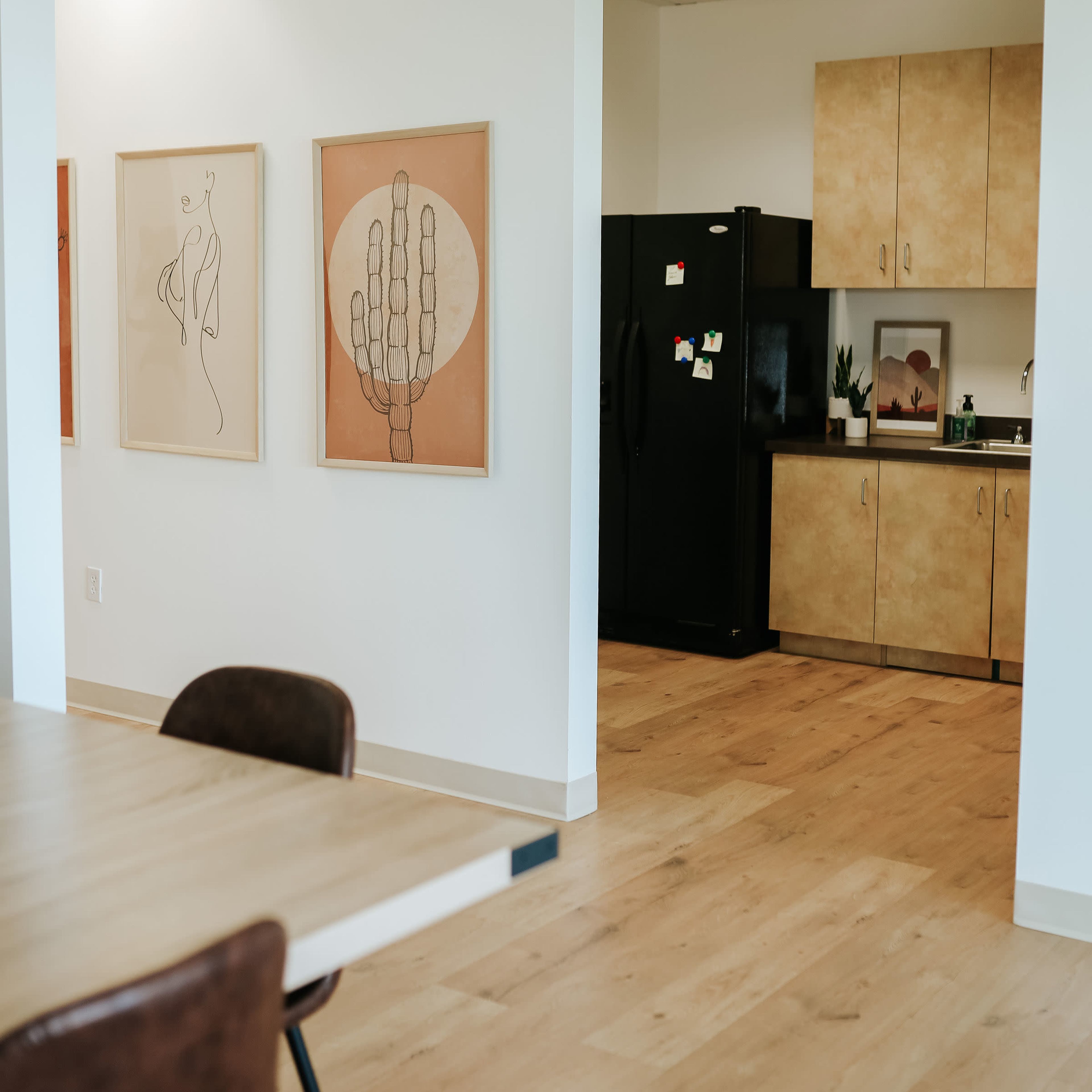 A modern interior features a dining area with a table and chairs, adjacent to a kitchen with a black refrigerator and artwork on the walls.