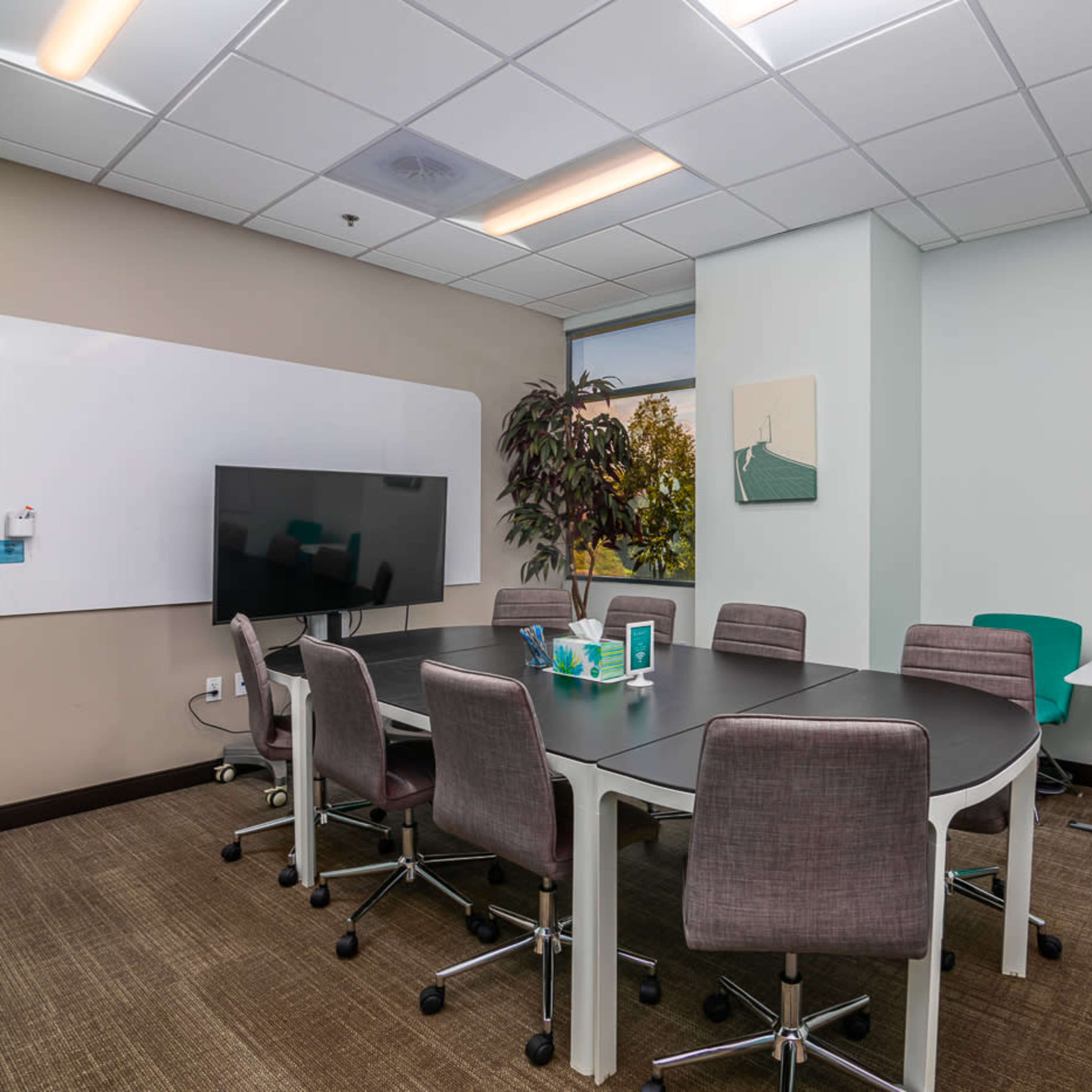 A conference room features a round table surrounded by chairs, a large screen, and a whiteboard on the wall.