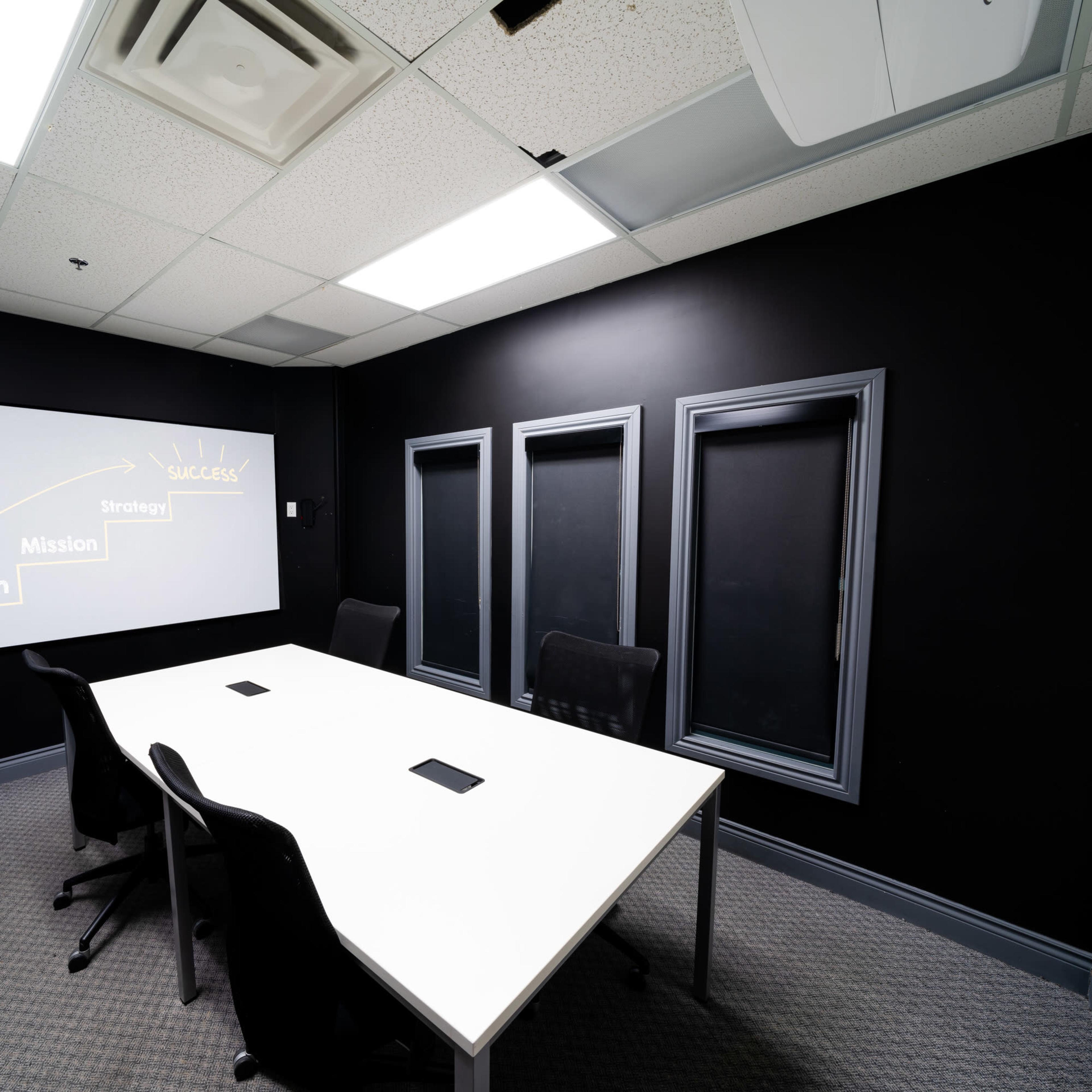 A conference room features a white table surrounded by black chairs, with a projector screen displaying strategic goals on a dark wall.