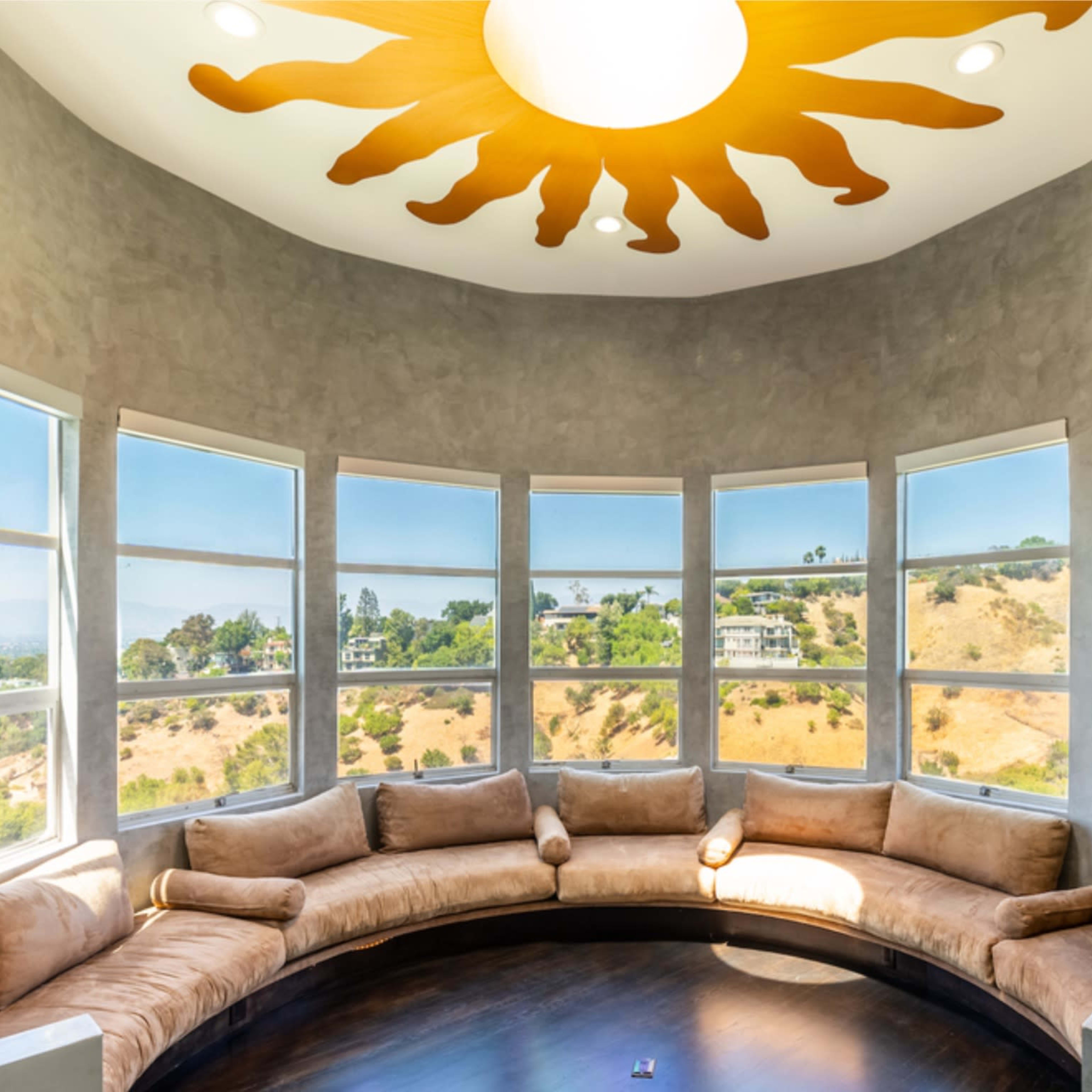 A rounded seating area with a sun-shaped ceiling light, surrounded by large windows that overlook a hilly landscape.
