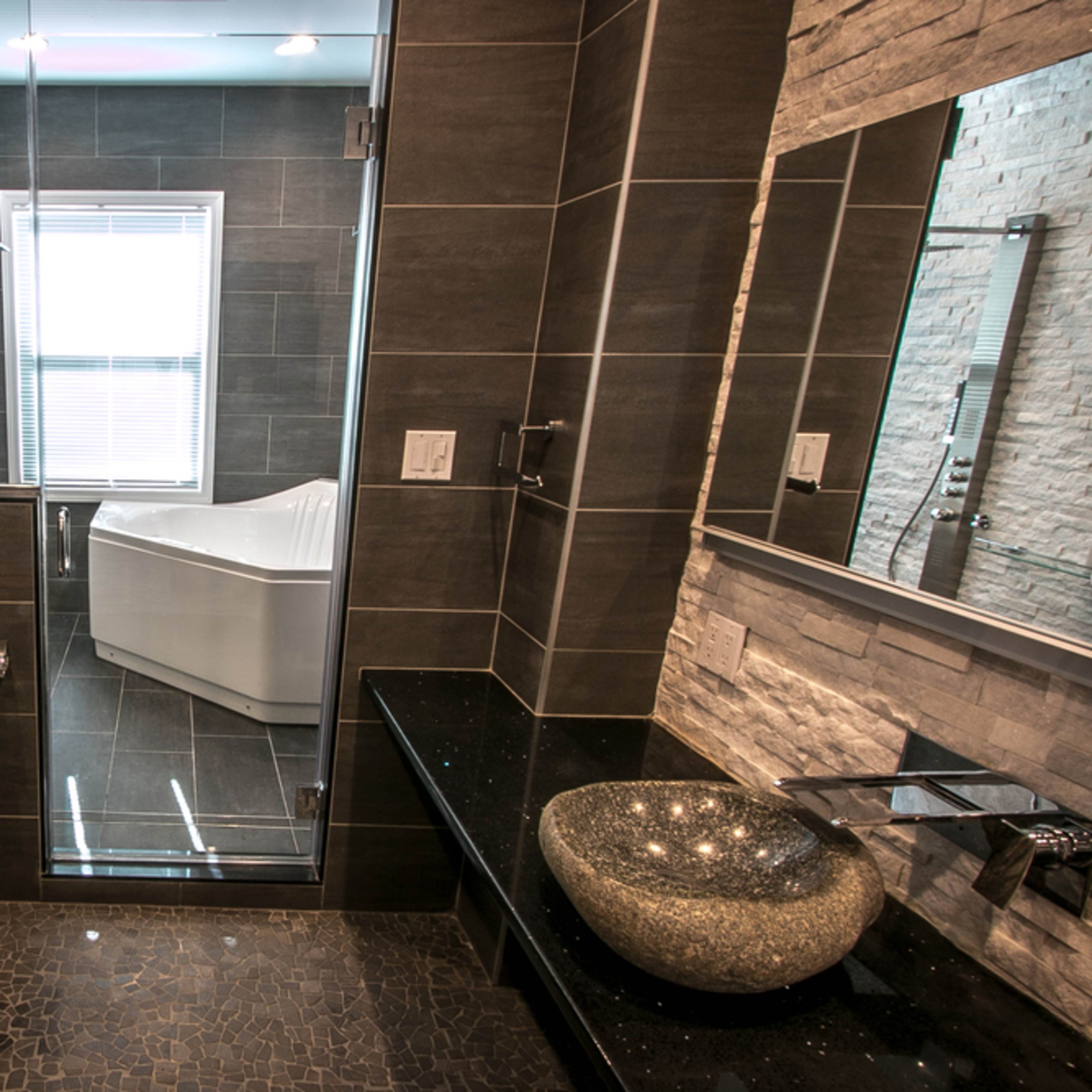 A modern bathroom featuring a glass shower enclosure, a freestanding bathtub, a sleek toilet, a stone sink with a faucet, and dark tiled walls.