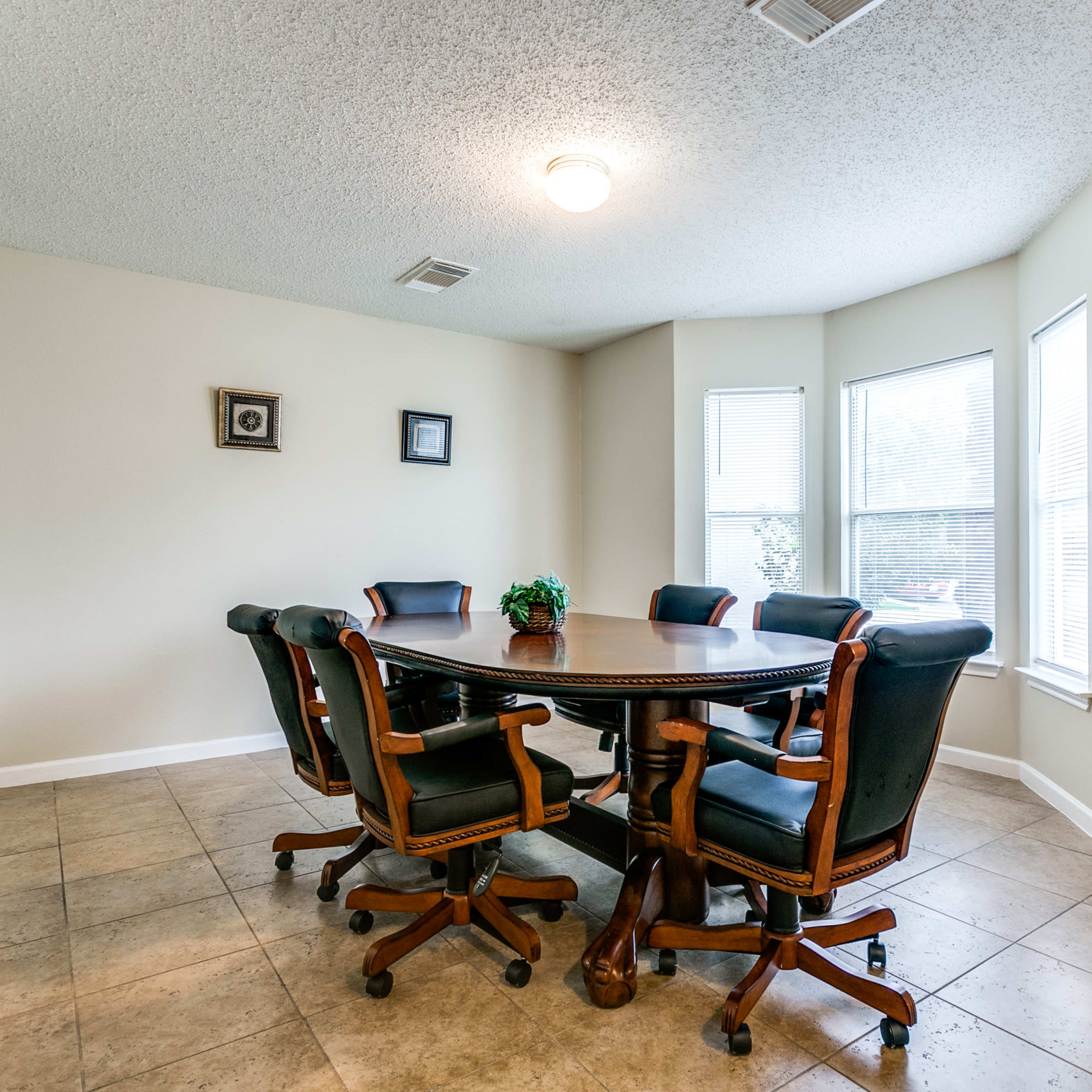 A round wooden table surrounded by six black and brown office chairs is set in a well-lit corner of a room with large windows.