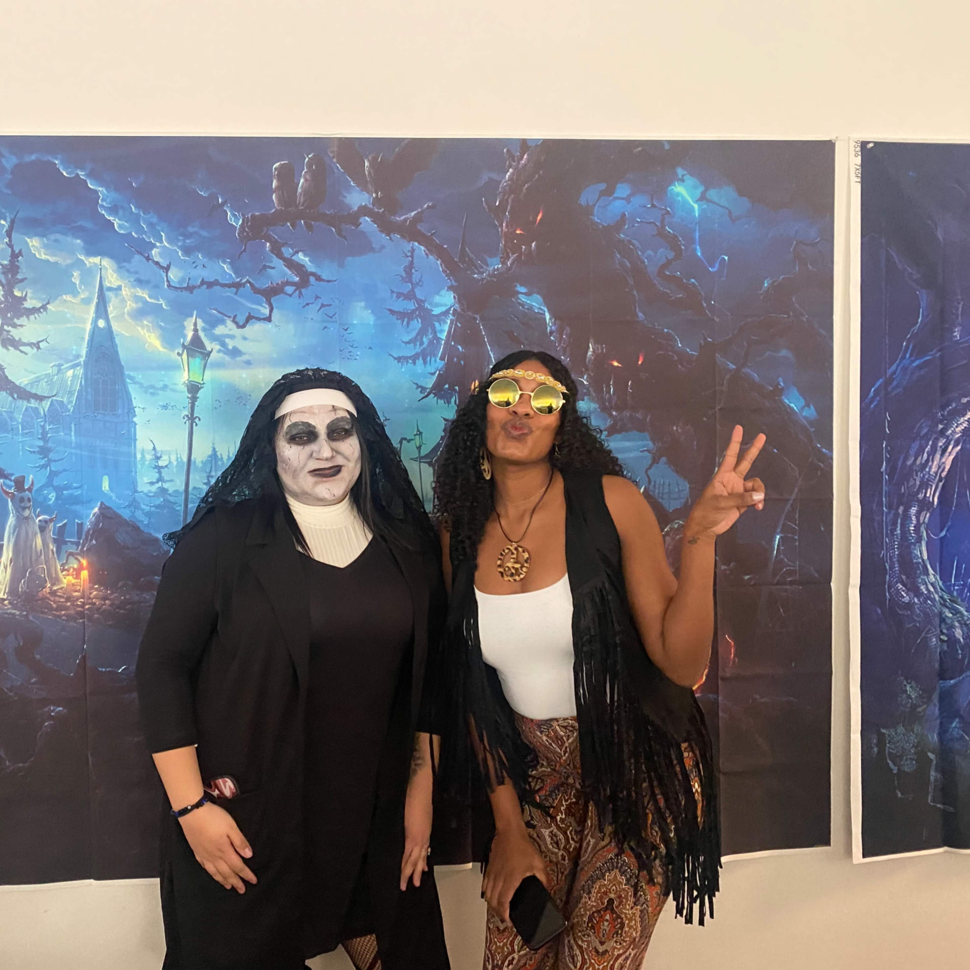 Two women pose for a photo in front of a large, dark-themed mural depicting a spooky landscape with a castle and trees.
