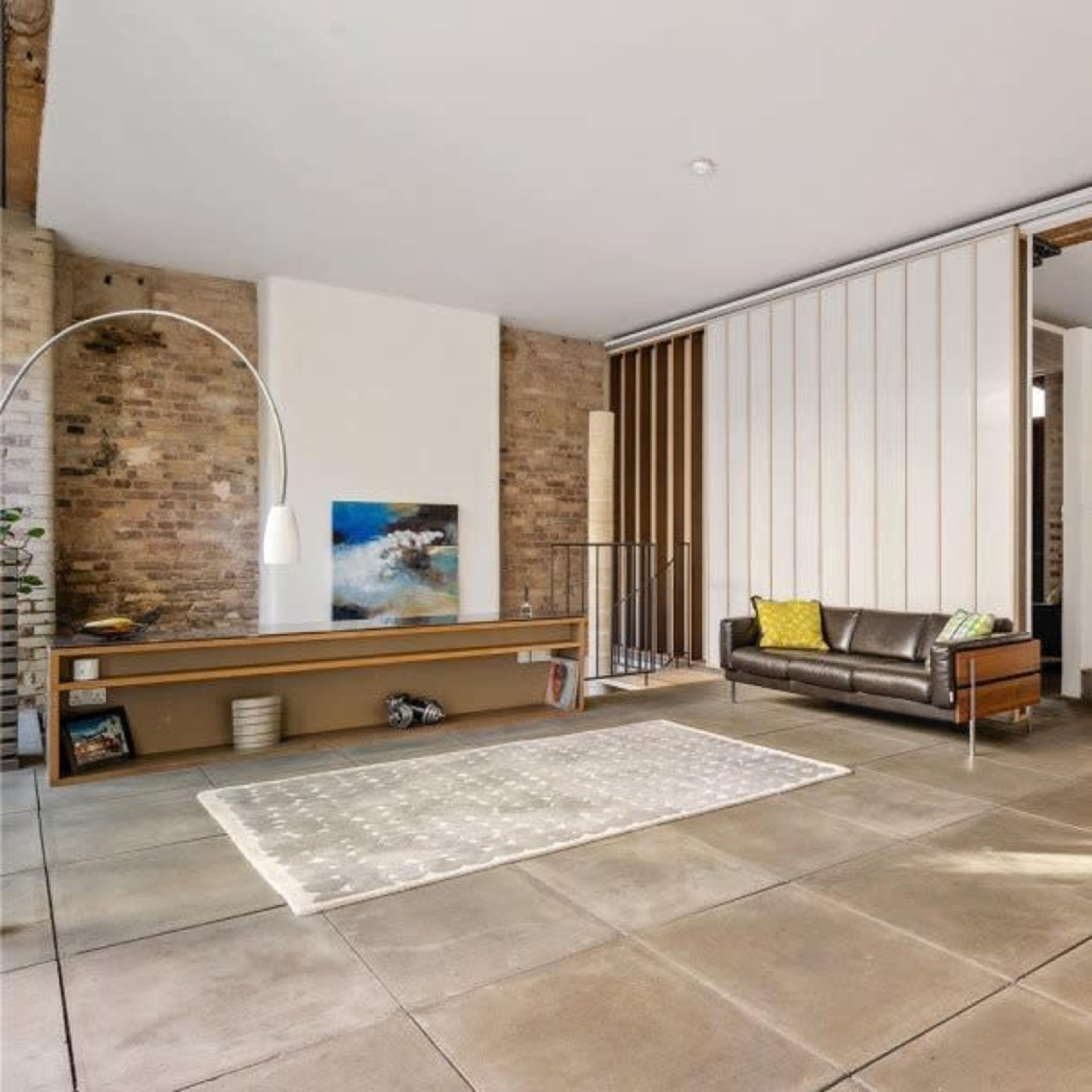 A modern living space featuring a leather sofa, a red egg chair, a patterned rug, and a staircase leading to an upper level, with exposed brick walls and large windows.