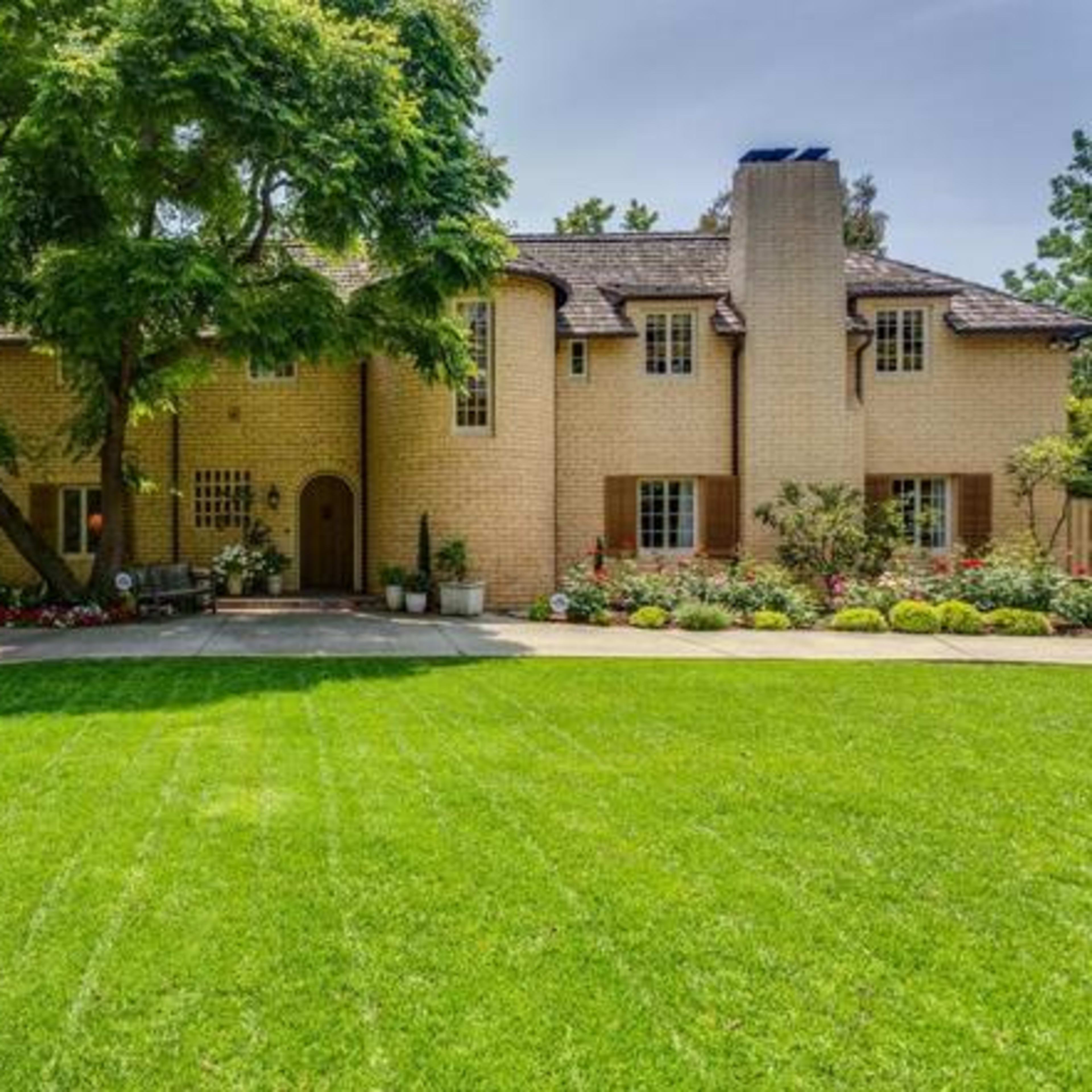 A large brick house with a chimney is set amidst well-maintained lawns and garden beds filled with flowers.