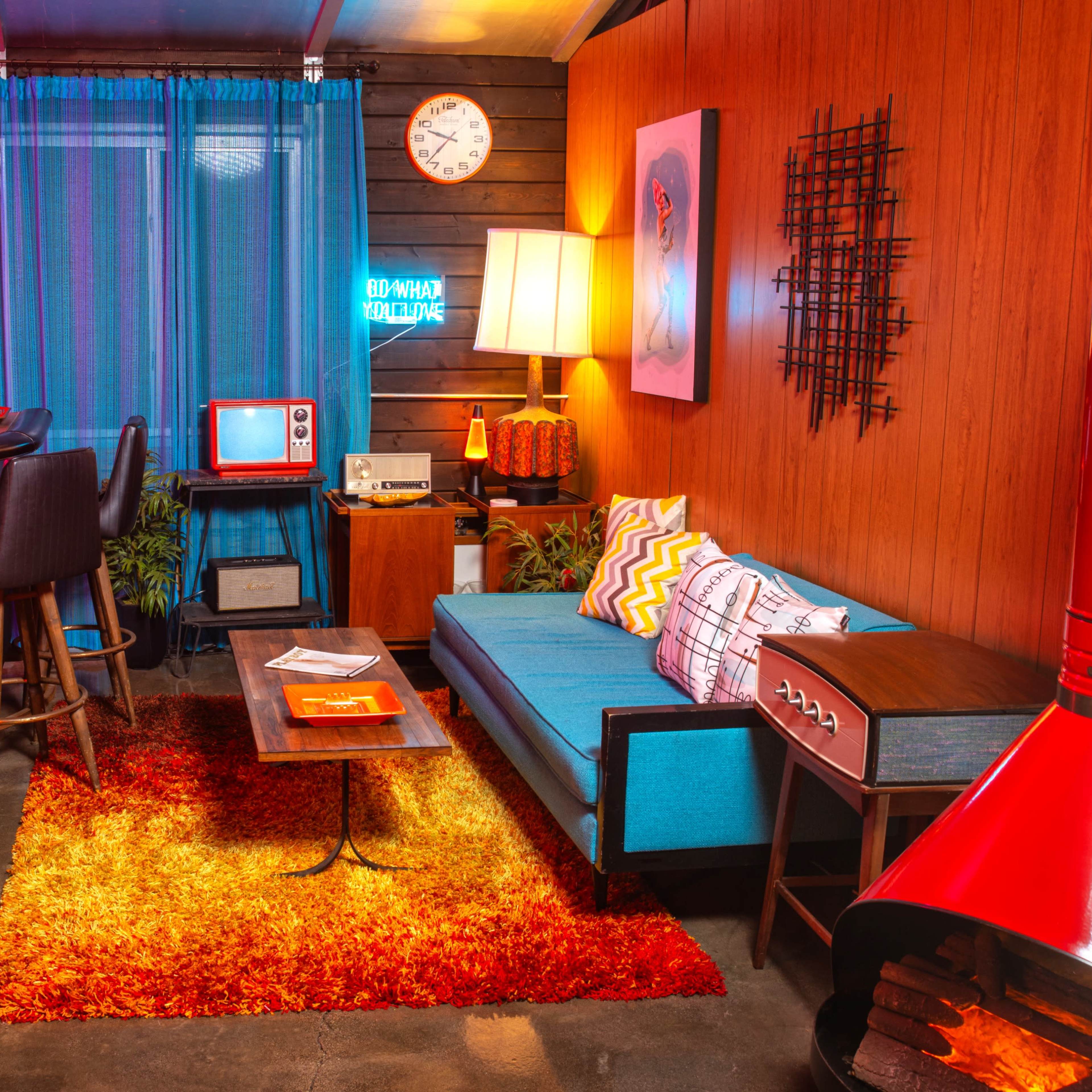 A mid-century modern living room with a blue sofa, a wooden coffee table, a bar area with stools, and colorful decor, including a retro television and a wall clock.