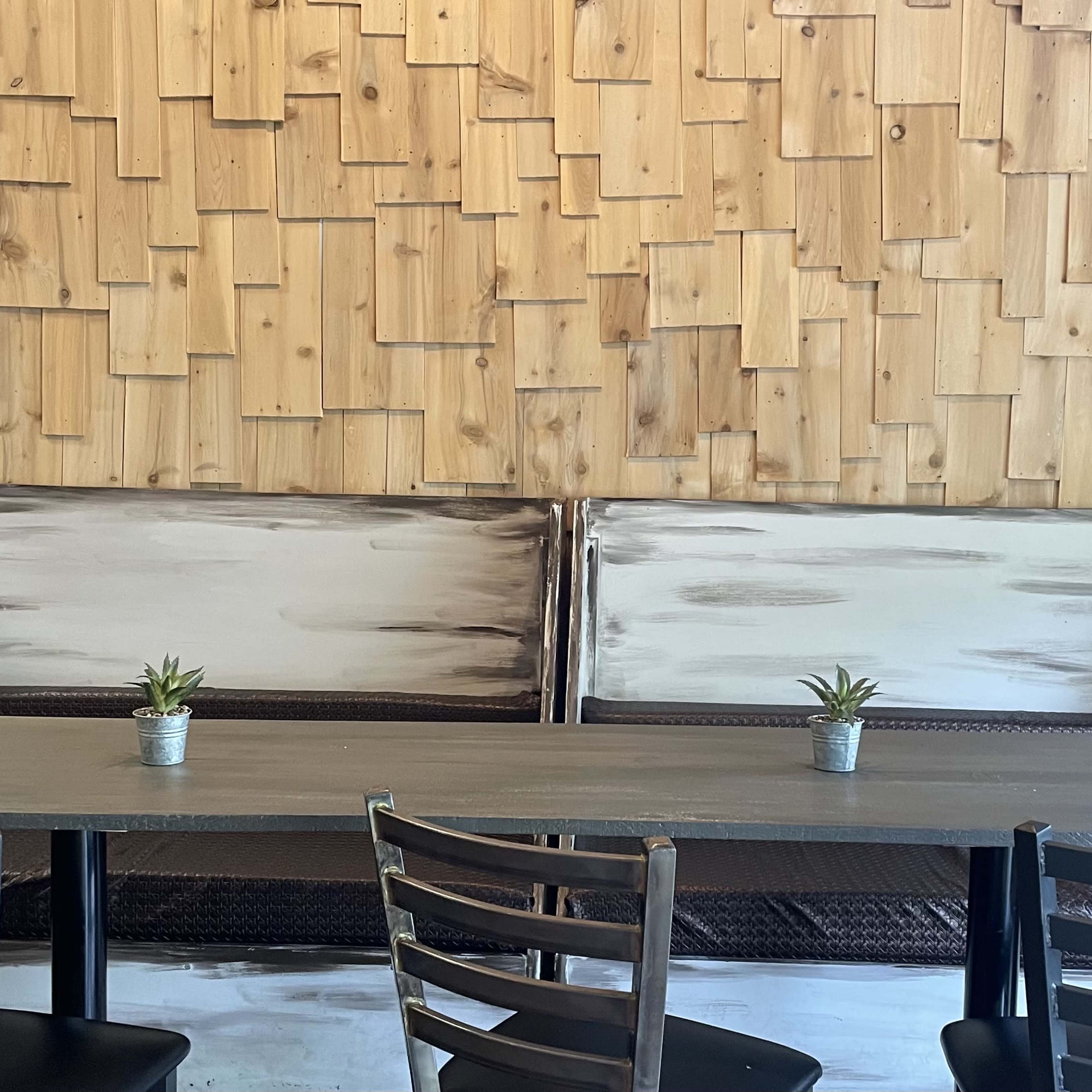 A seating area in a cafe with two wooden benches against a textured wooden wall and a table in front, each with a small potted plant on top.
