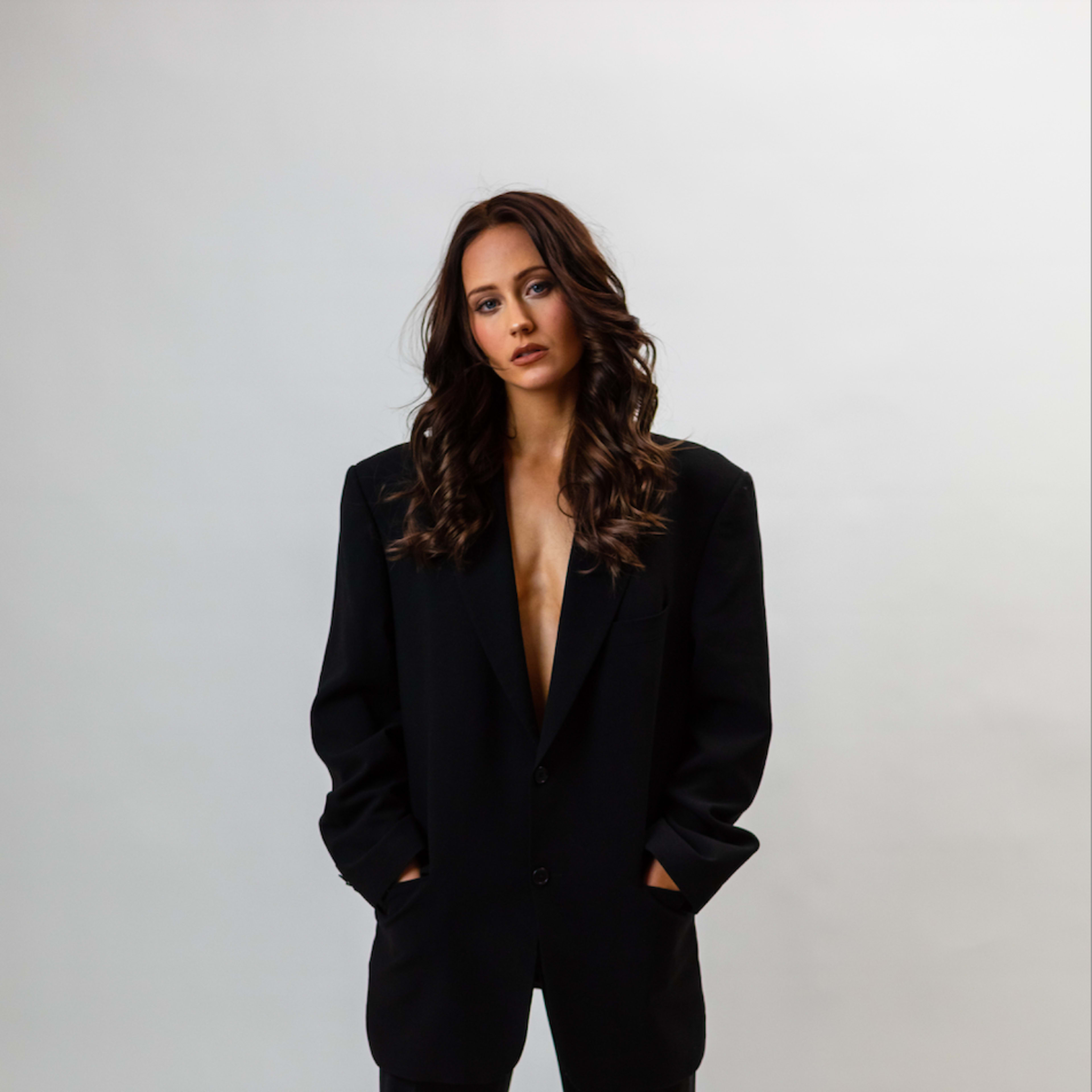 A person stands confidently in a black oversized suit with a deep neckline, hands in pockets, against a plain white background.