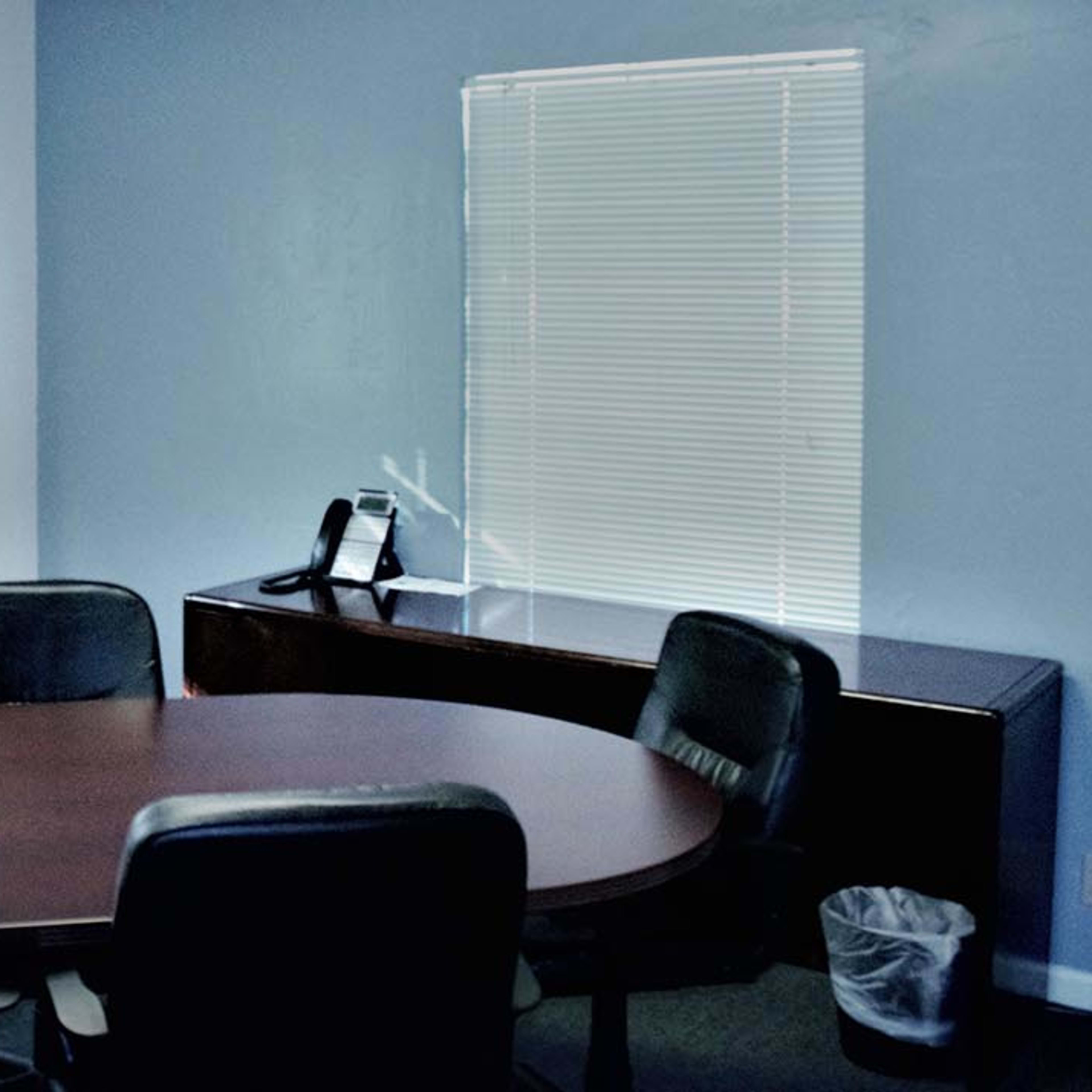 A conference room features an oval wooden table surrounded by leather chairs, with blue walls and blinds-covered windows.
