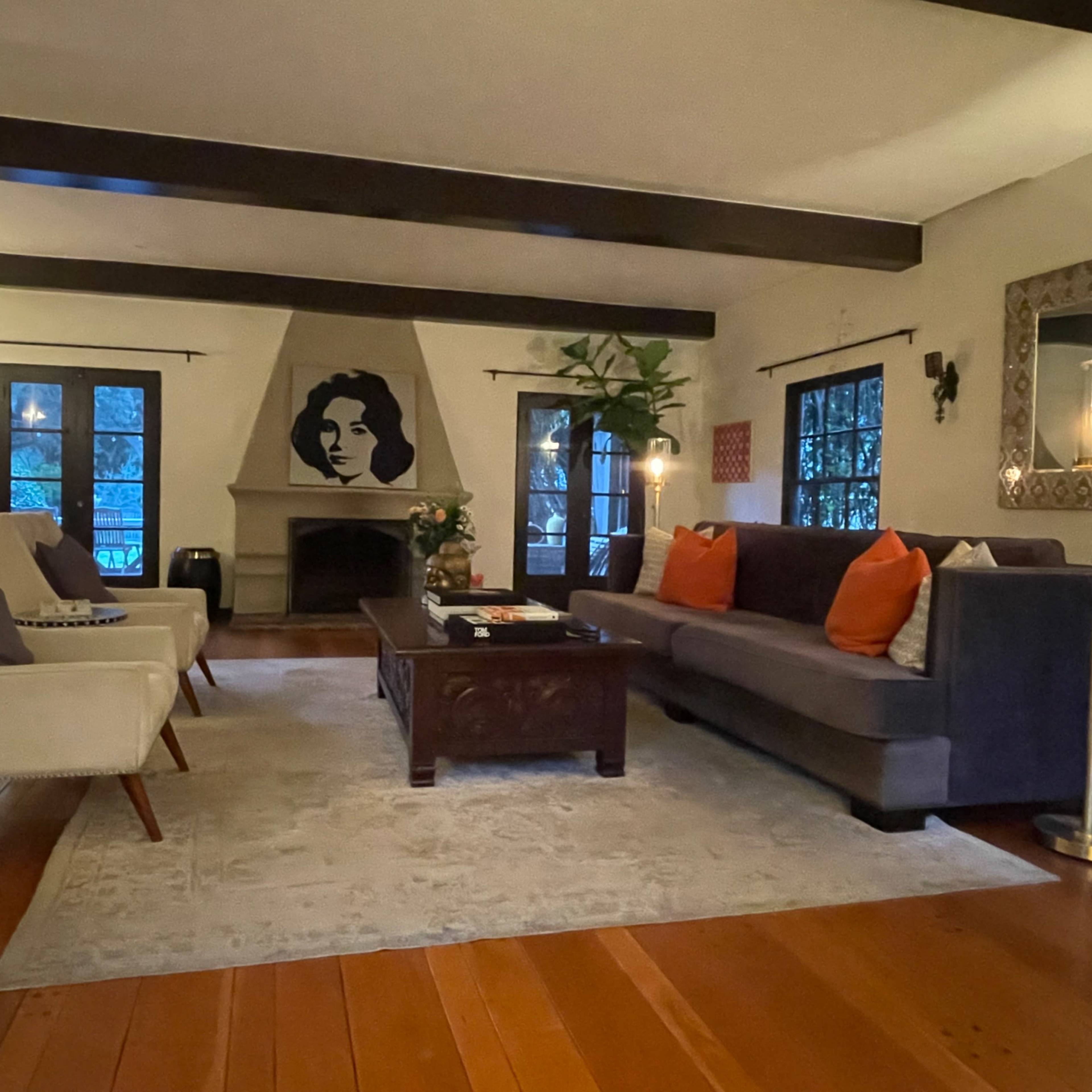 The living room features a dark sofa adorned with orange pillows, a decorative area rug on wooden flooring, a wall mural of a woman's face, and large windows framed by black curtains.