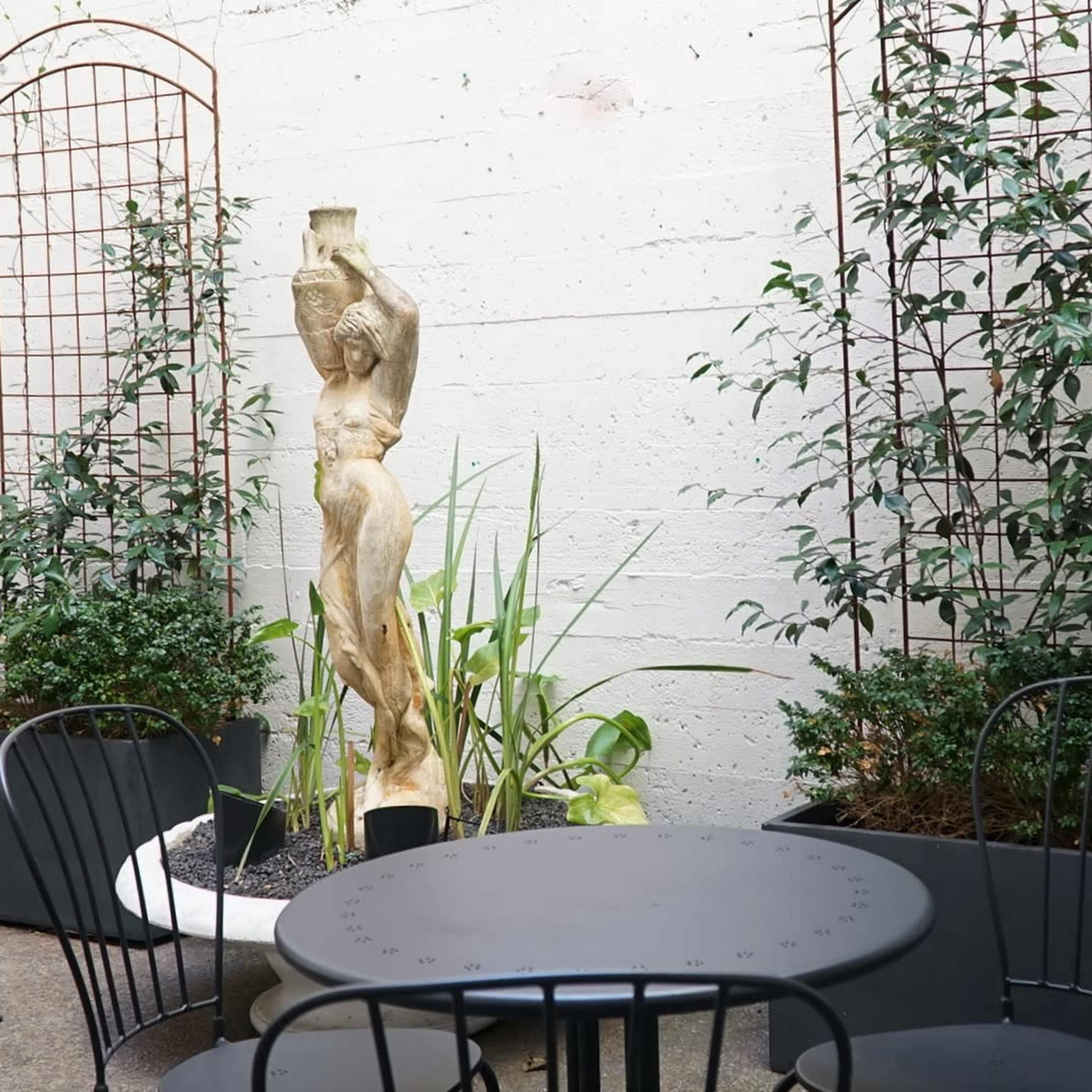 A courtyard features a round black table and chairs surrounded by greenery, including planters and trellises, with a sculpture of a figure in the center.