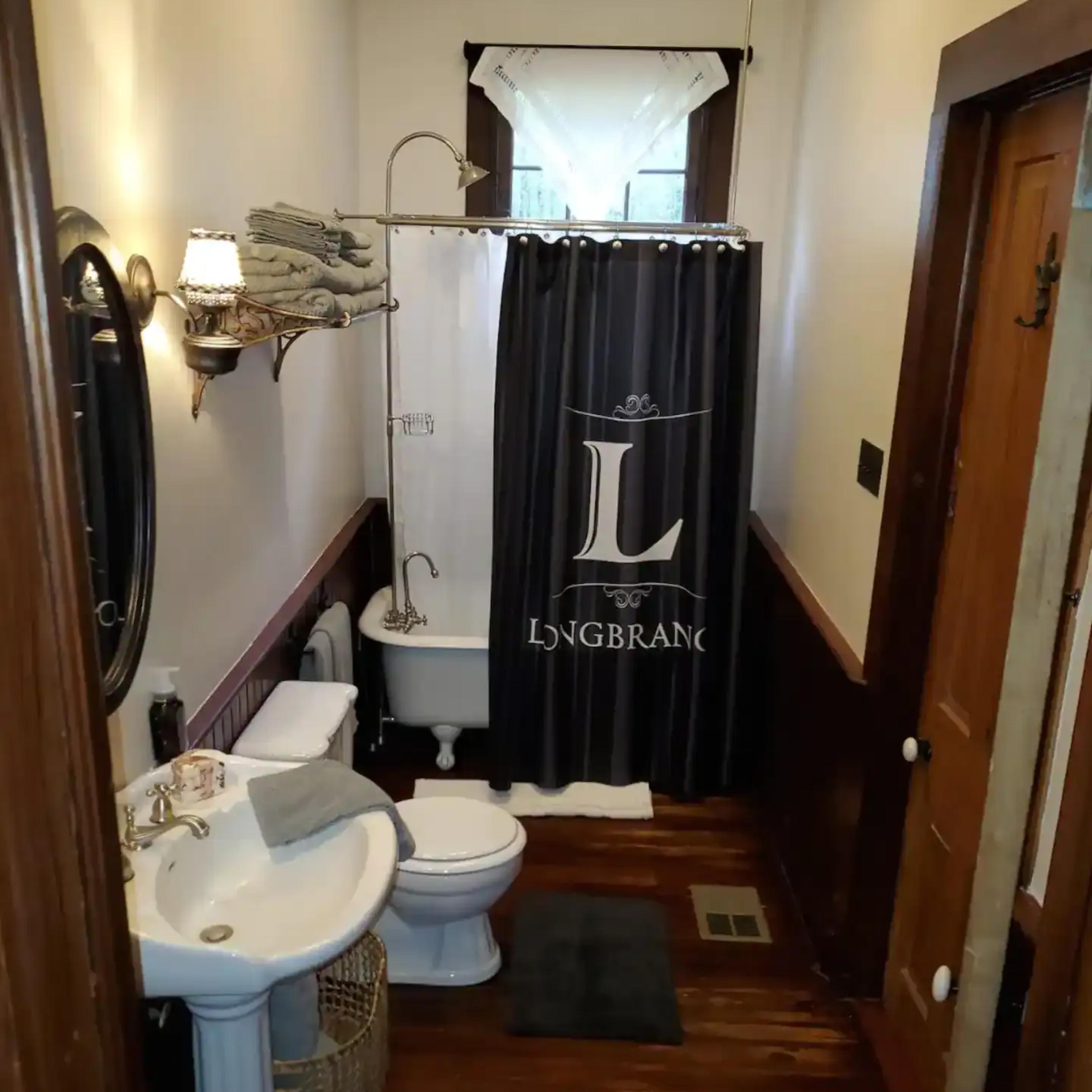 A small bathroom featuring a pedestal sink, a toilet, and a shower curtain with a logo, complemented by wooden floors and two rugs.