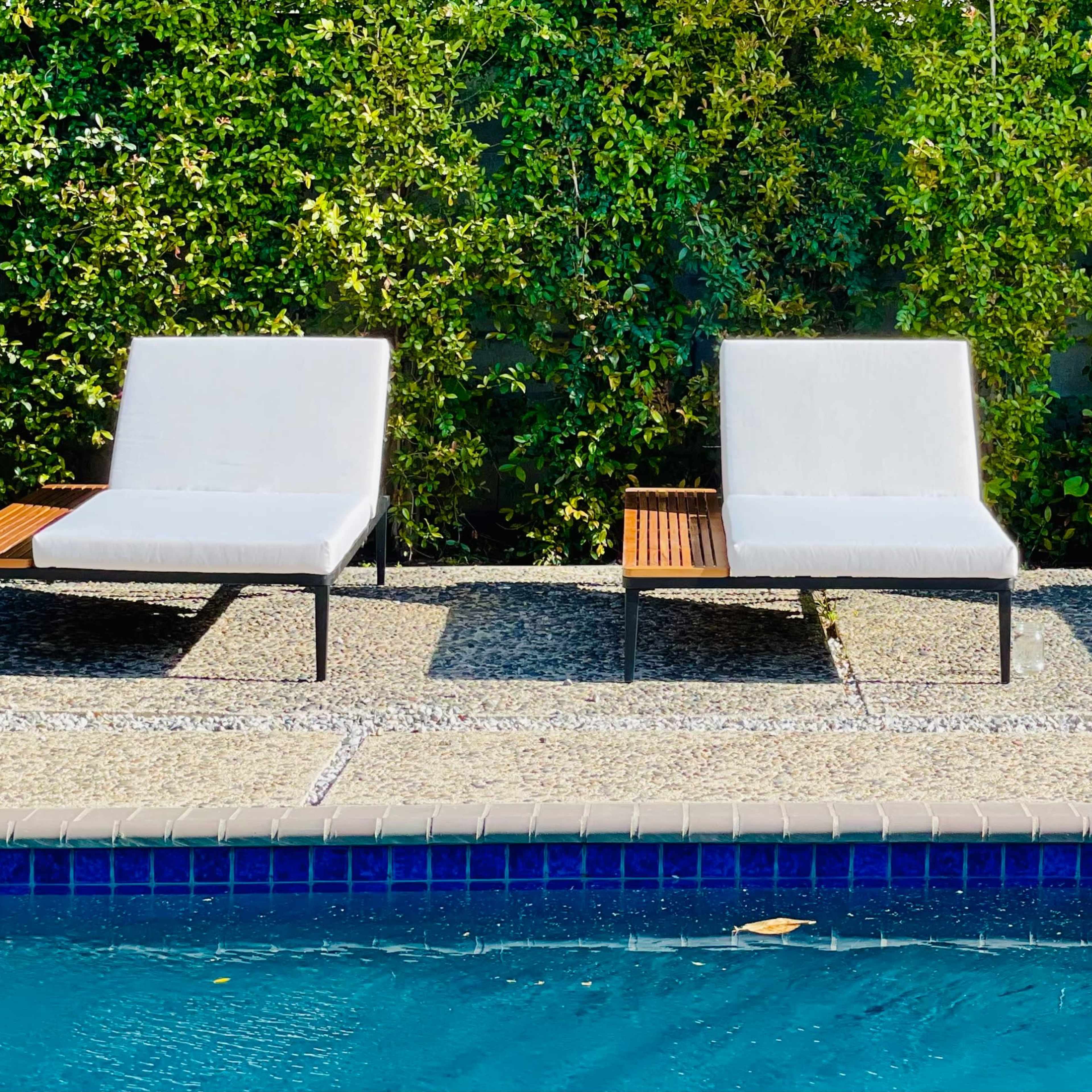 Three lounge chairs with wooden armrests are arranged beside a swimming pool, set against a backdrop of lush greenery.