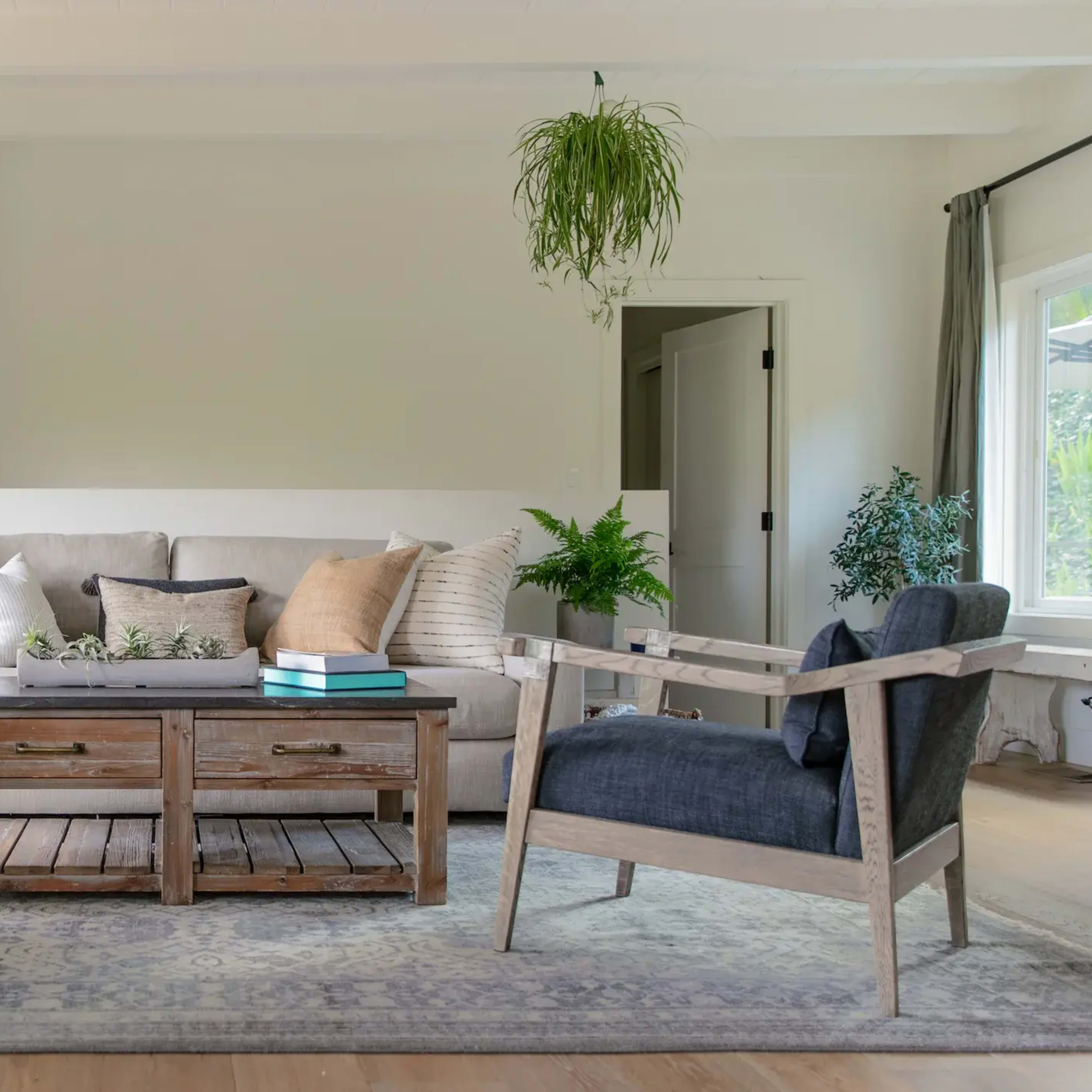 A living room with a beige sofa, two wooden armchairs, a coffee table made of reclaimed wood, and plants hanging from the ceiling.
