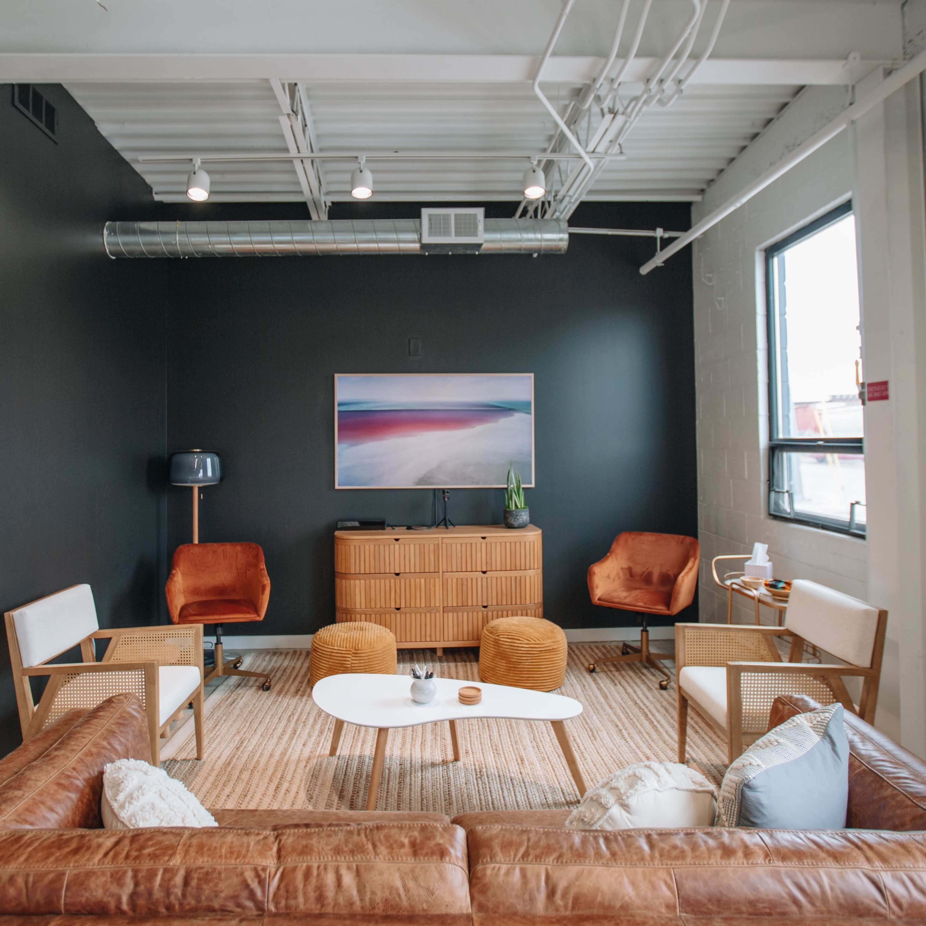 A modern lounge area with a brown leather sofa, a white coffee table, two orange armchairs, a wooden media console, and a large abstract painting on a black wall.