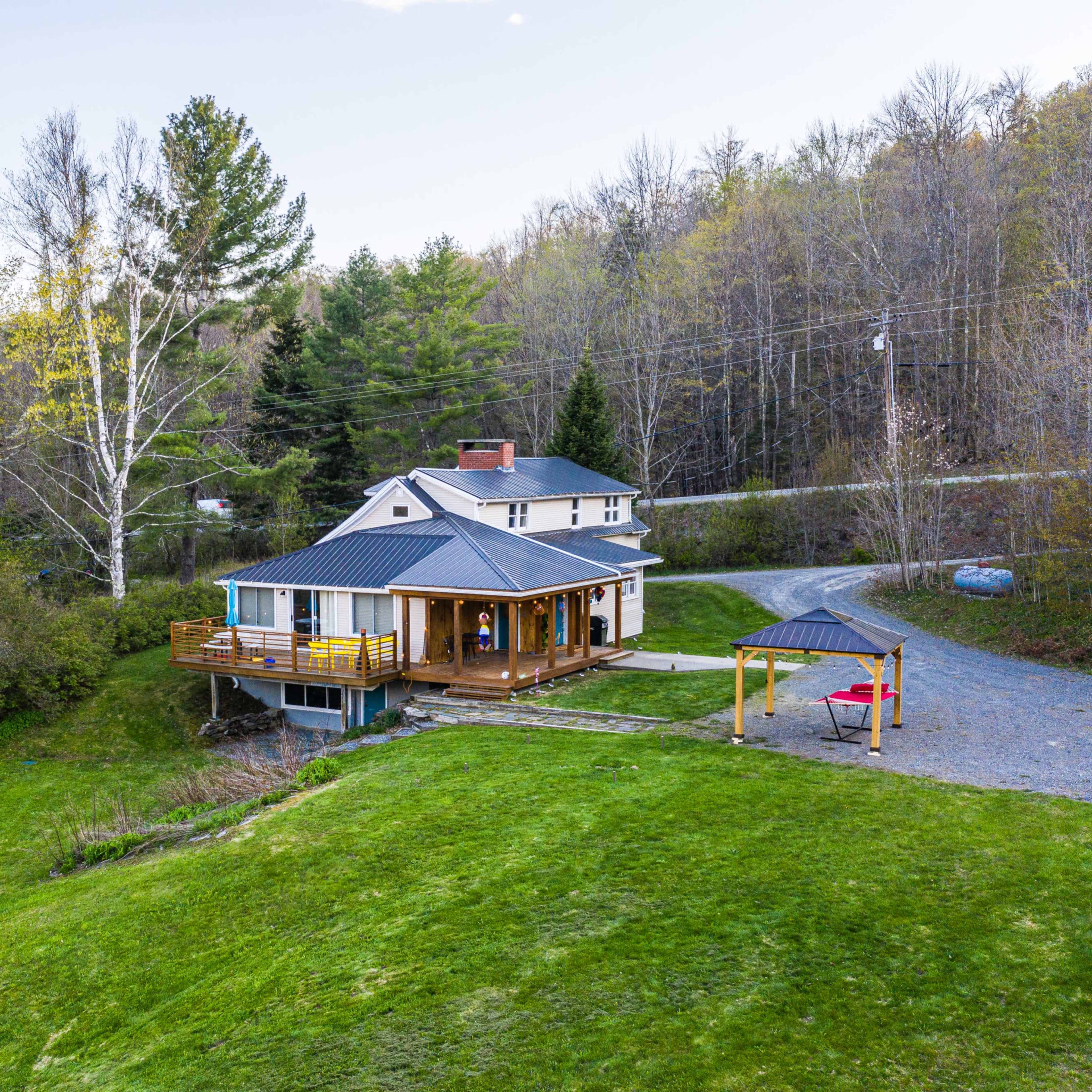 A two-story house with a wooden deck overlooks a grassy slope and is surrounded by trees.