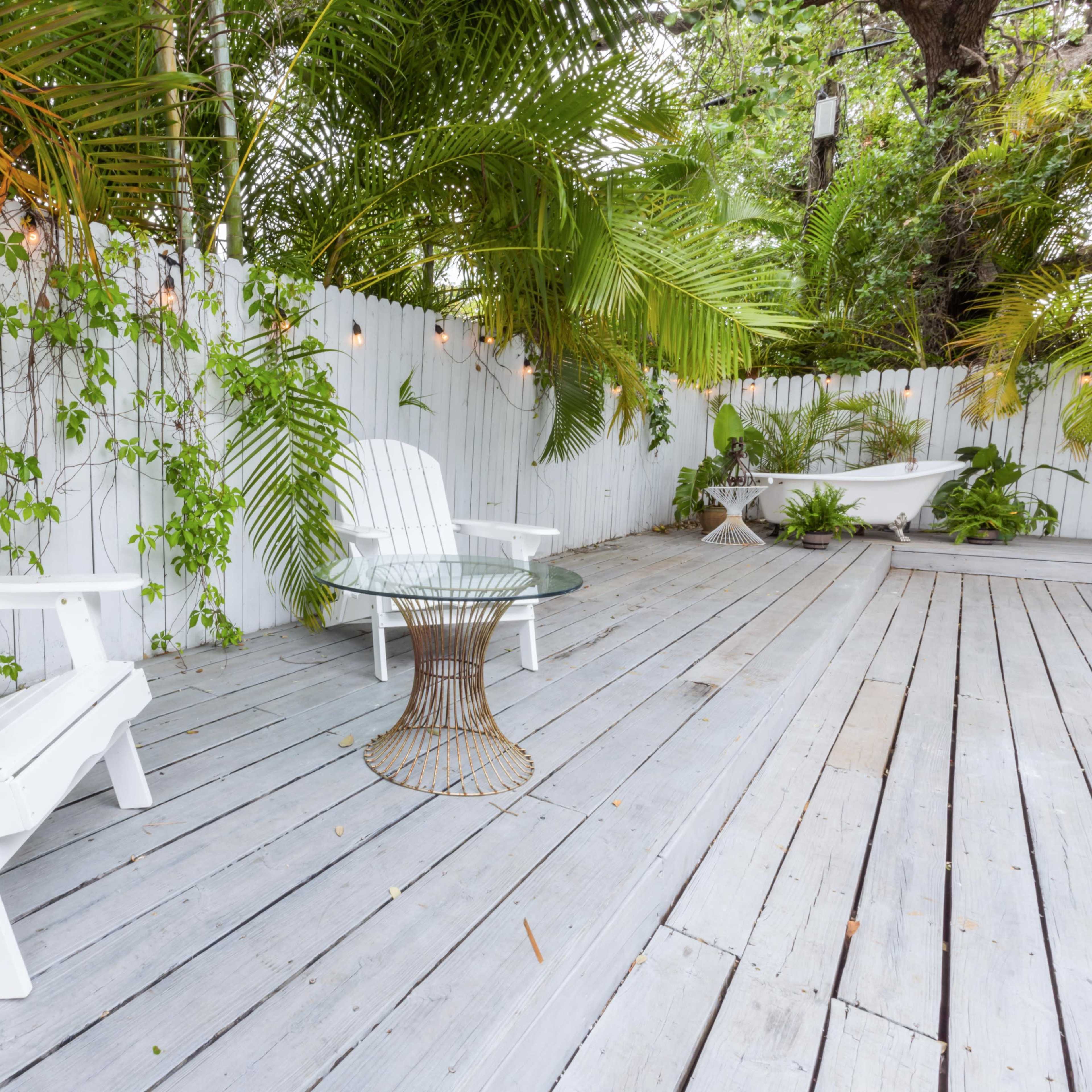 A pair of white Adirondack chairs and a glass table are set on a wooden deck surrounded by lush greenery and a white fenced wall.