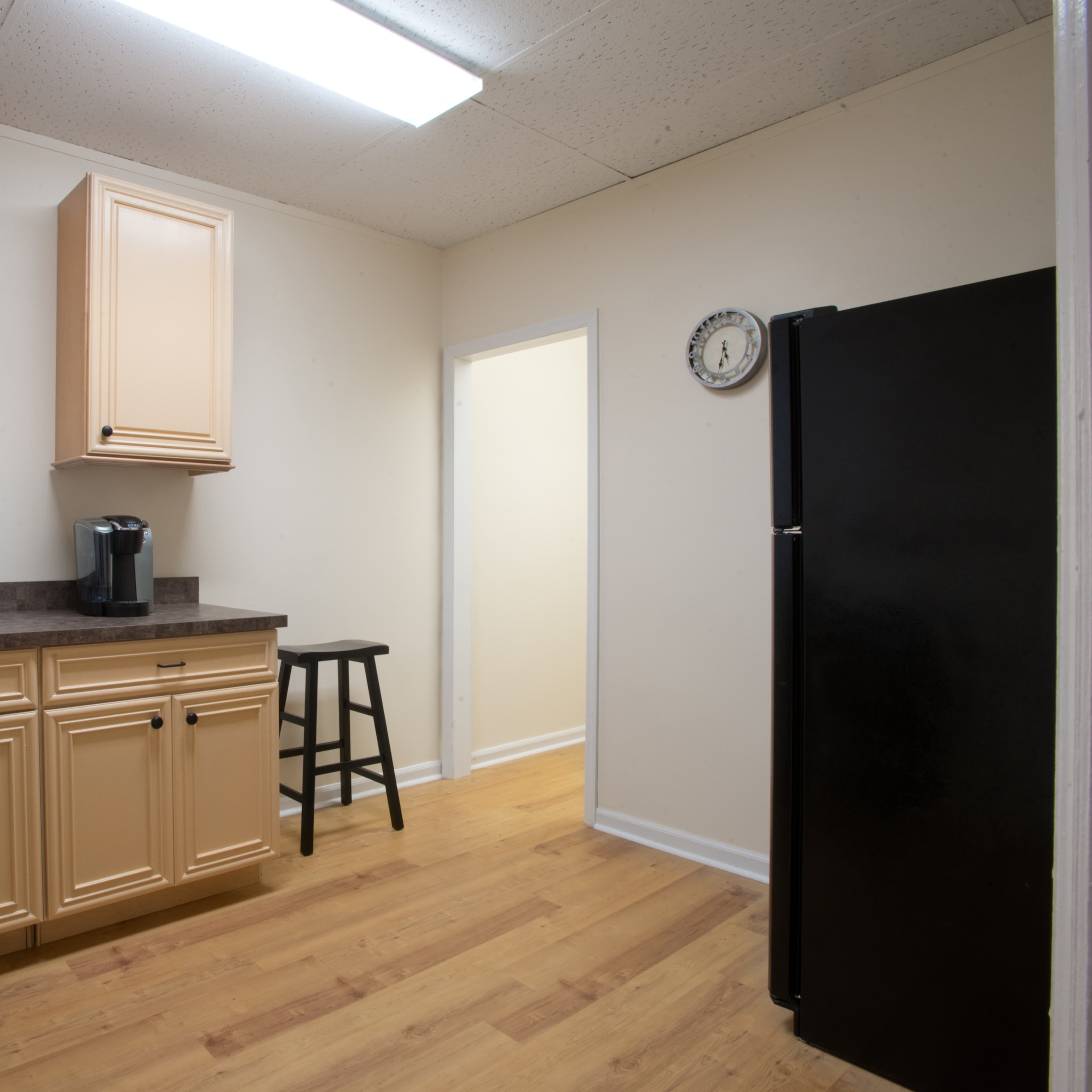 A small kitchen with light-colored cabinets, a dark countertop, a coffee maker, a black refrigerator, and a single stool.