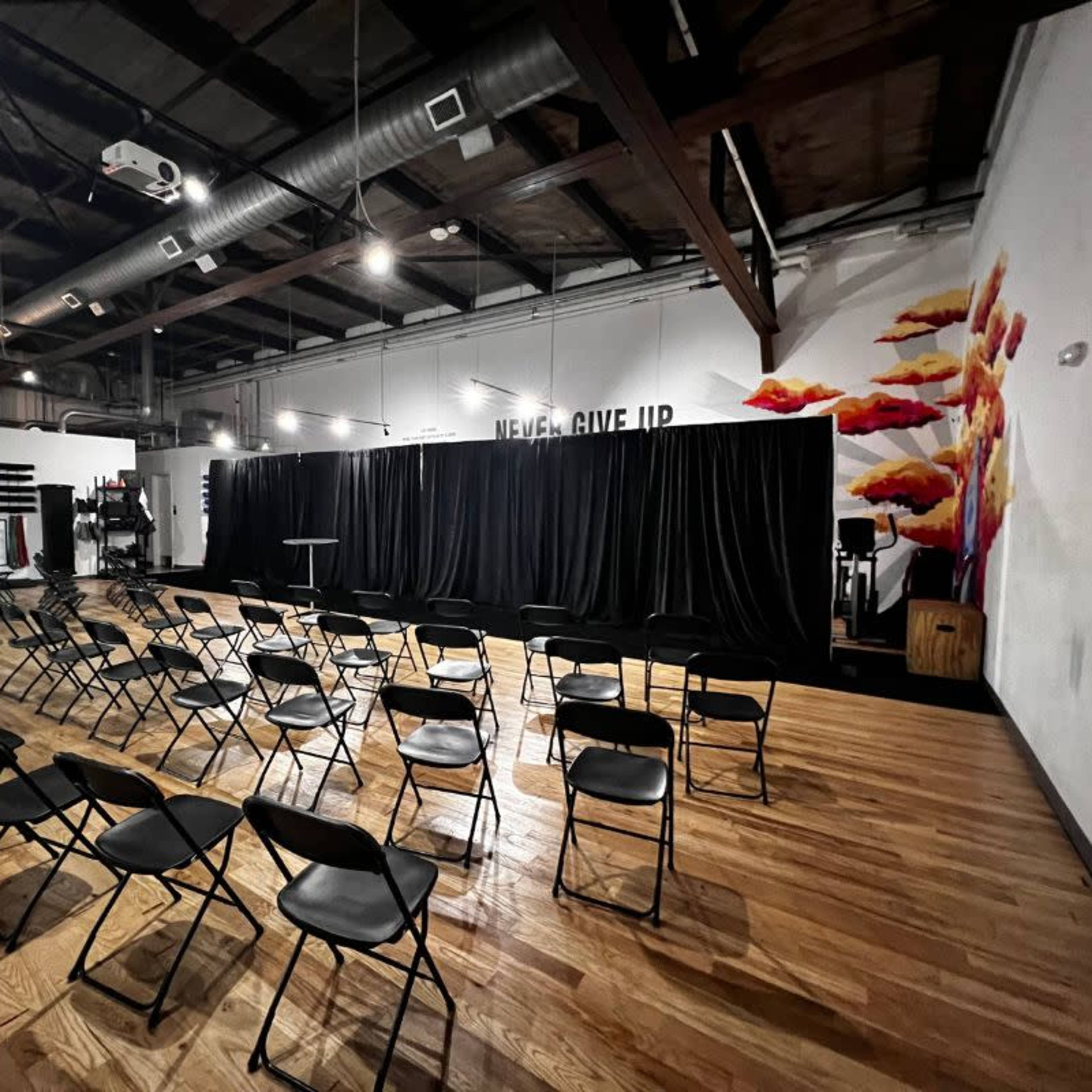 A spacious room set up with rows of black chairs facing a black curtain stage, with motivational text on the wall and a colorful mural.