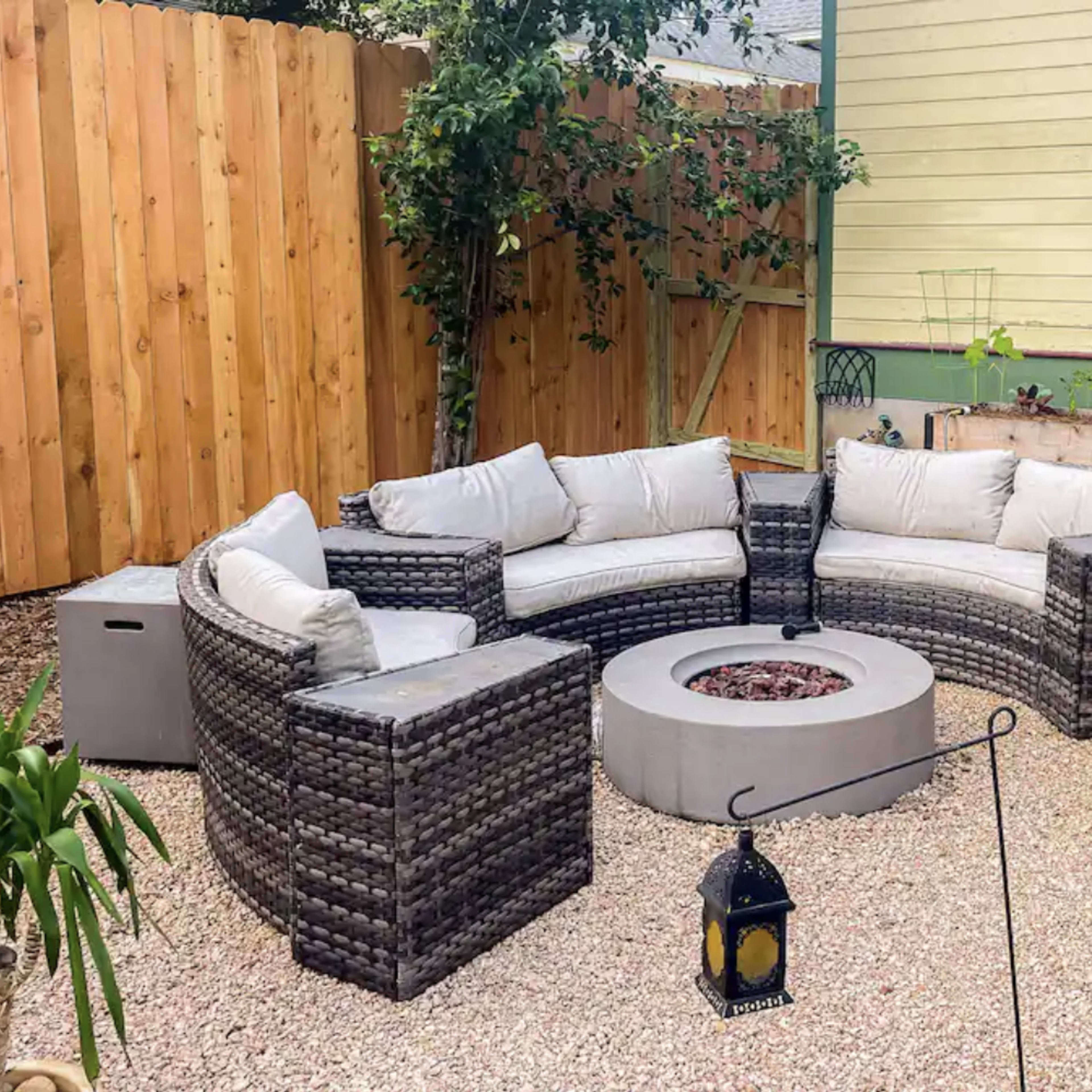 A patio area with a circular fire pit surrounded by a curved seating arrangement made of wicker furniture, set among gravel and various plants.