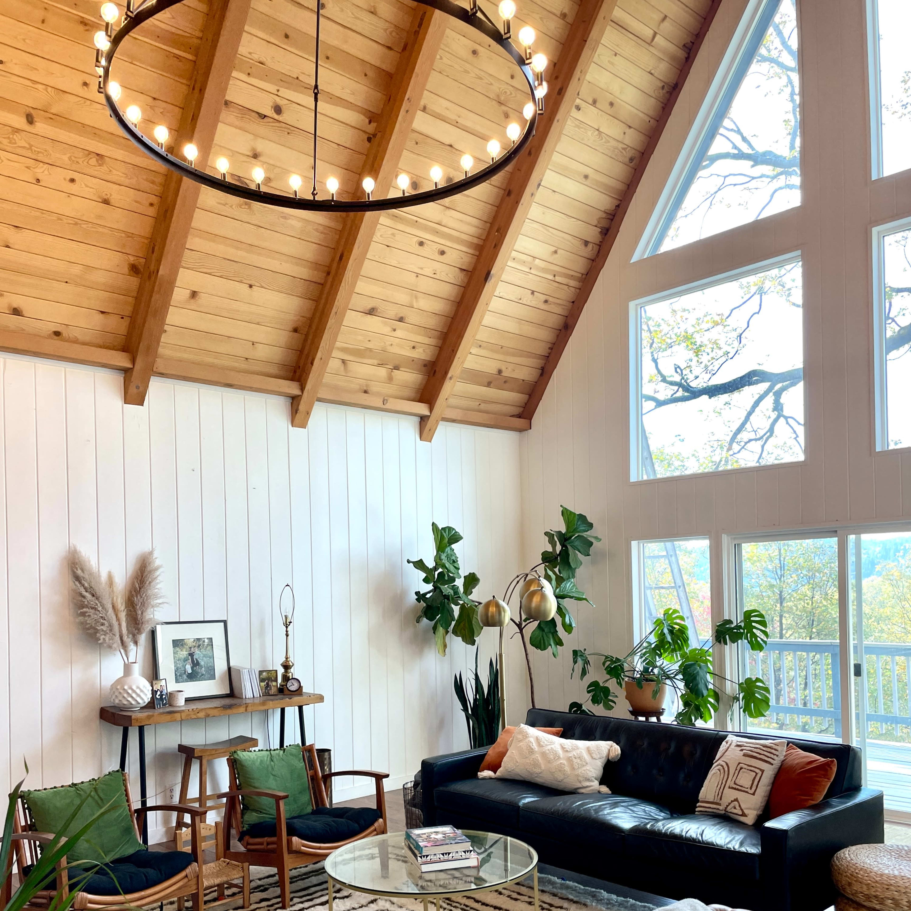 A cozy living room with a high wooden ceiling, large windows, a black leather sofa, and a glass coffee table surrounded by plants and decorative elements.