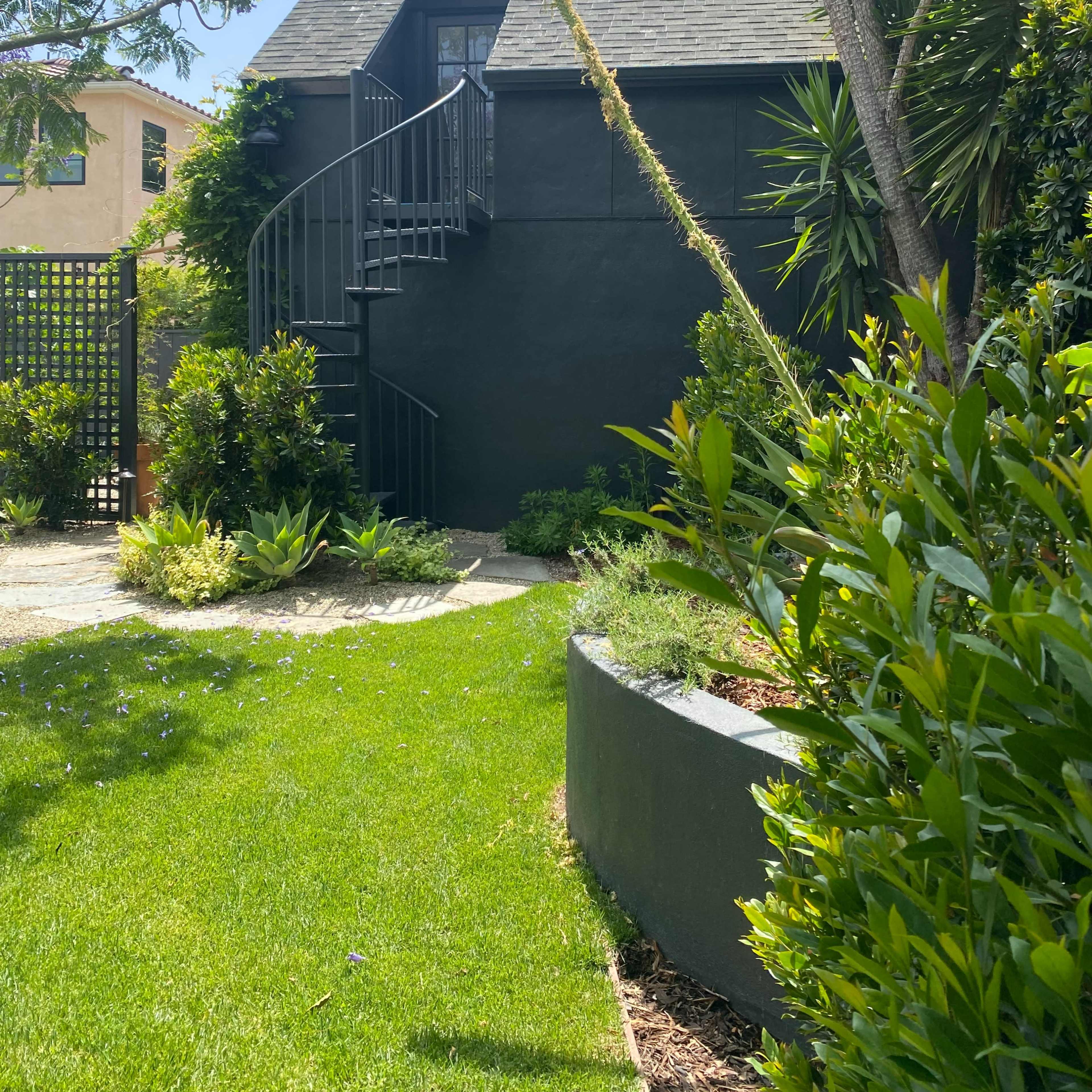 A black two-story house with a spiral staircase is surrounded by a landscaped garden and various plants.