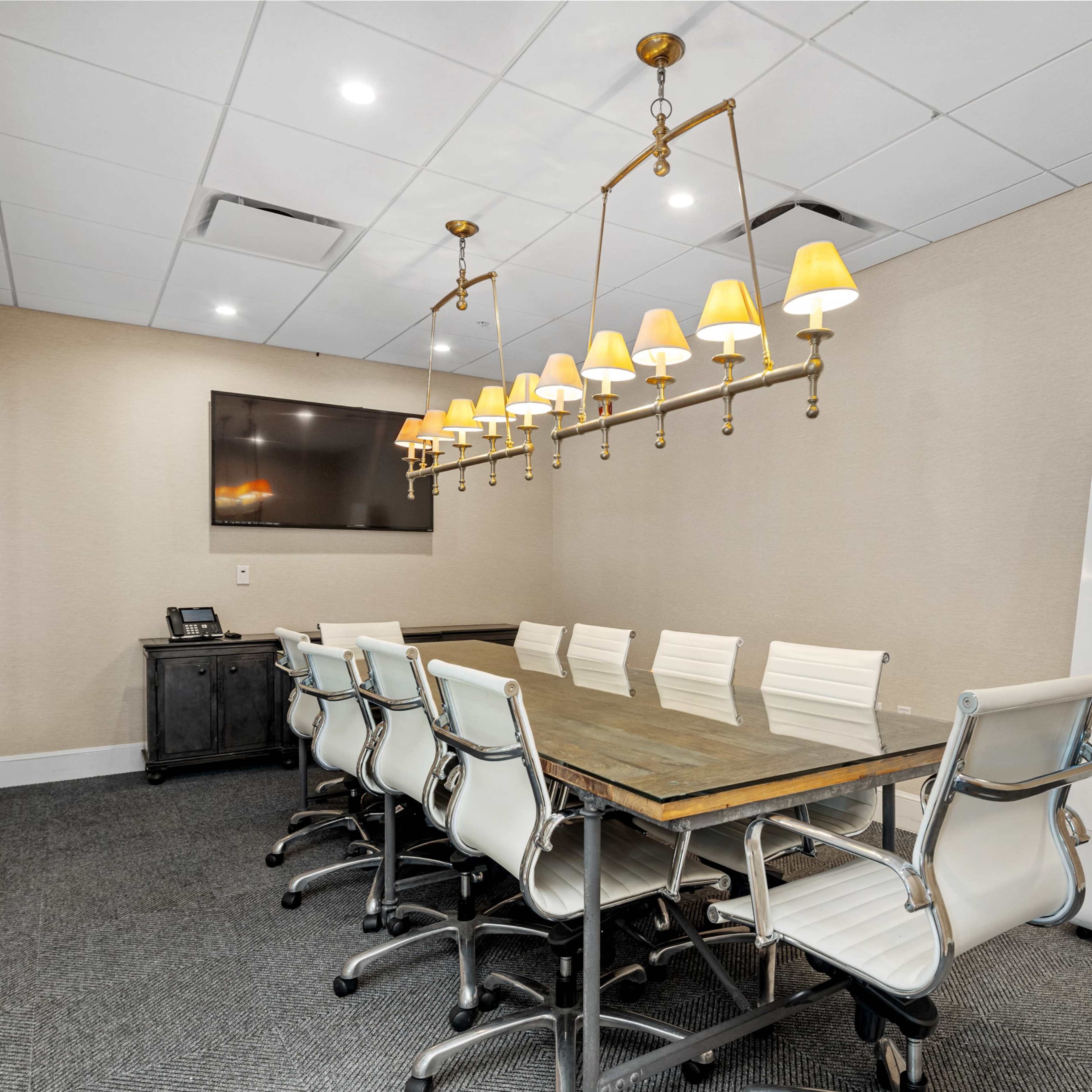 A conference room features a long wooden table surrounded by white chairs, with a row of pendant lights above and a wall-mounted screen on one side.