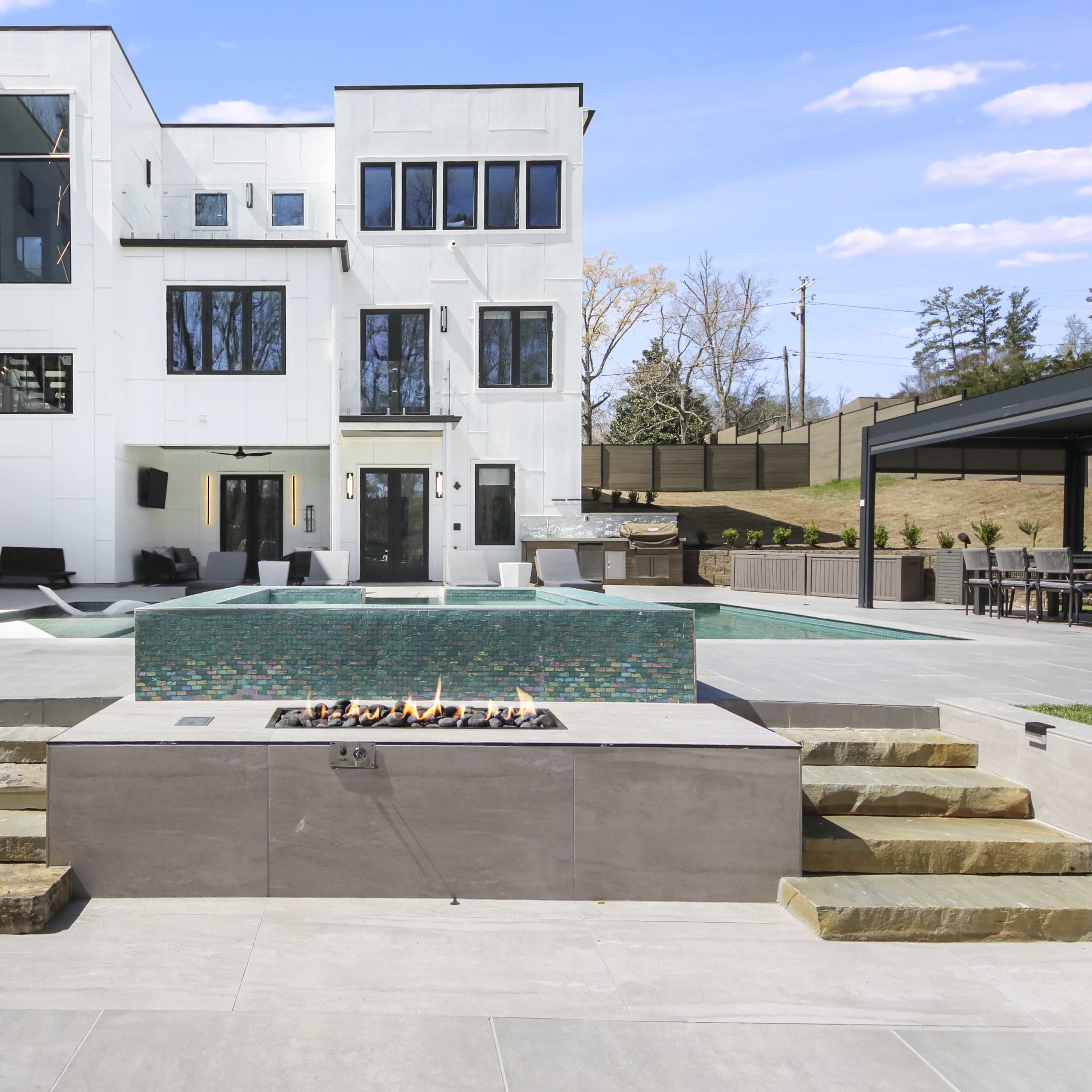 A modern outdoor patio area featuring a swimming pool, a fire pit with a stone surround, and a contemporary house in the background.