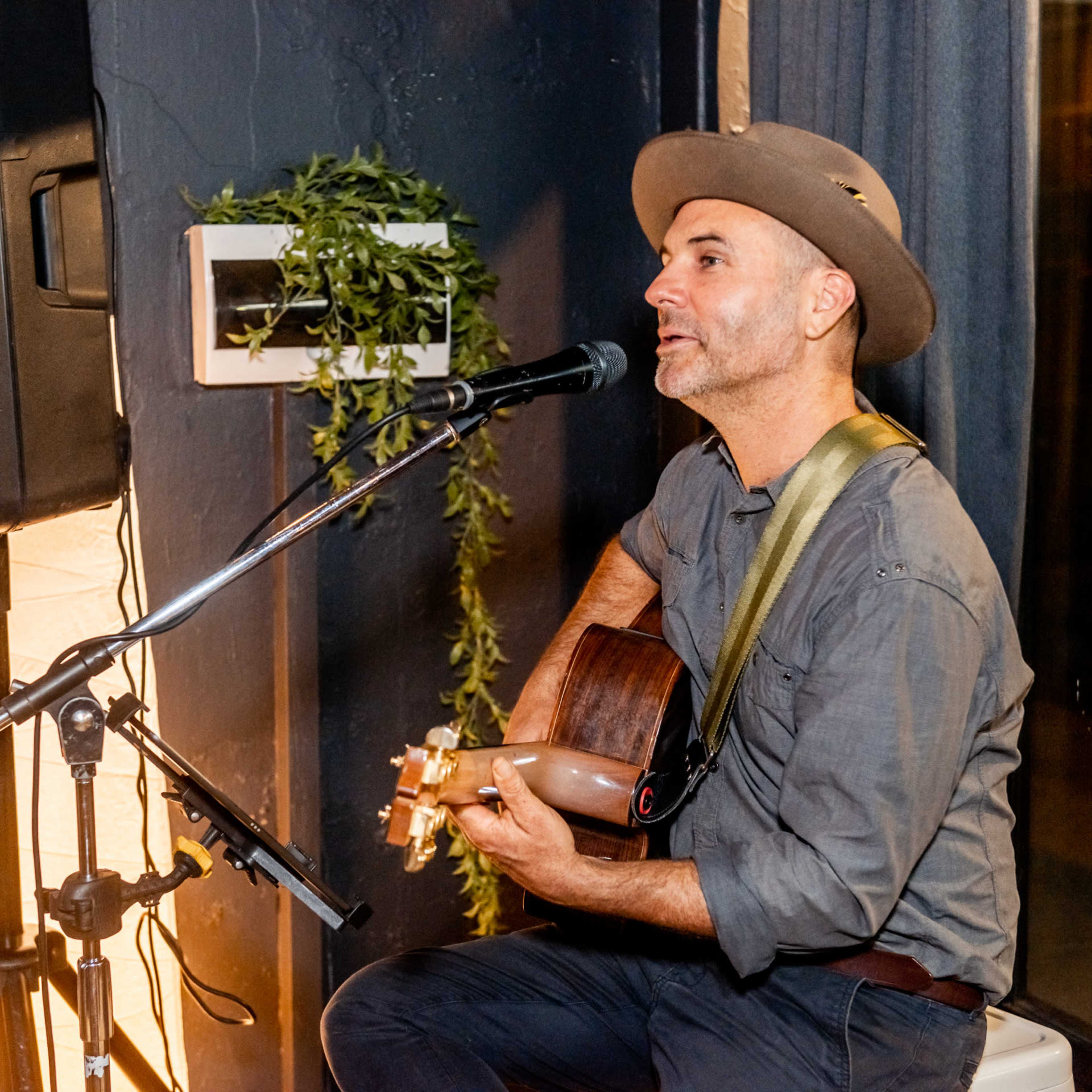 A man plays an acoustic guitar and sings into a microphone while seated next to a music speaker.