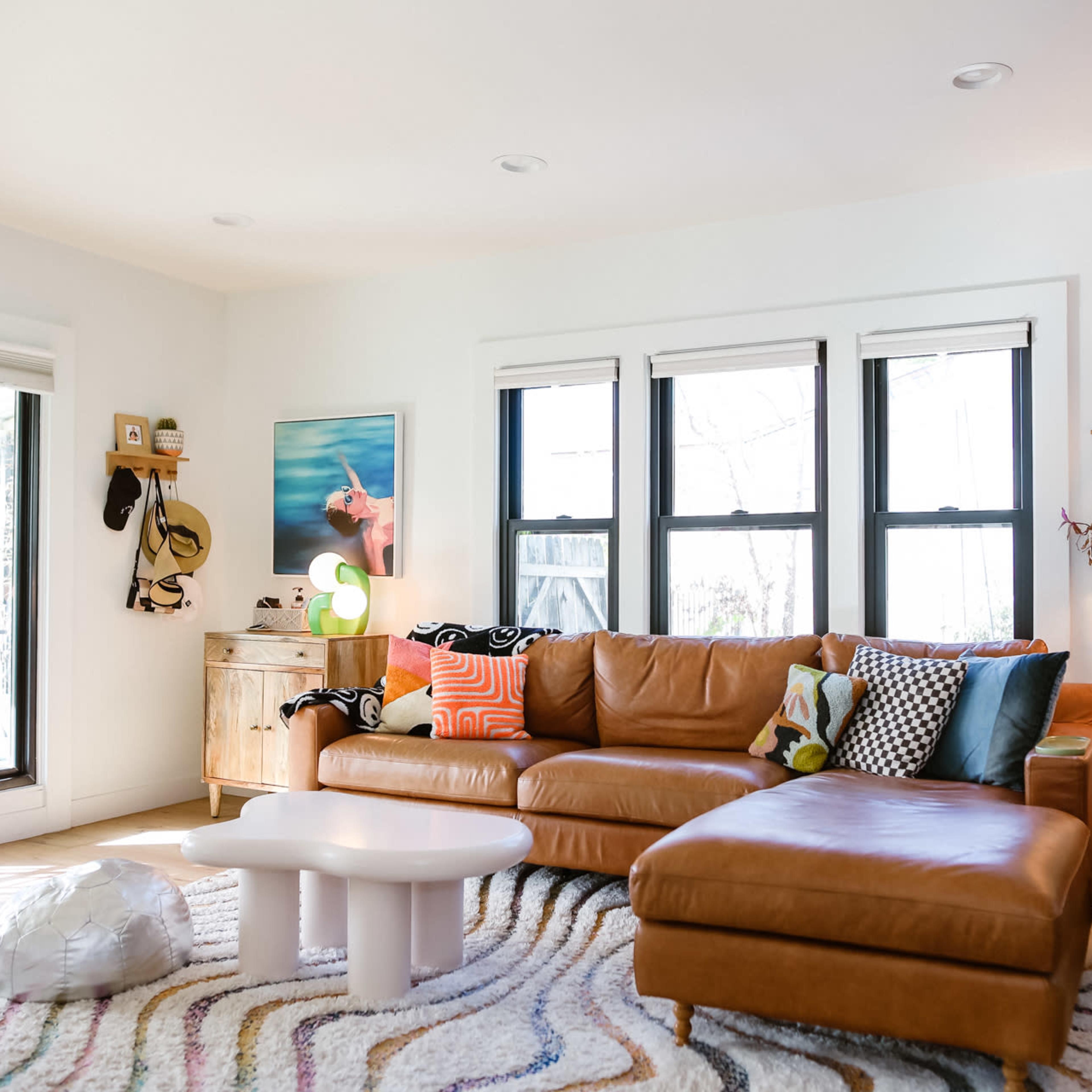 The living room features a brown leather sectional sofa, a white coffee table, and vibrant decor, with large windows allowing natural light to enter.
